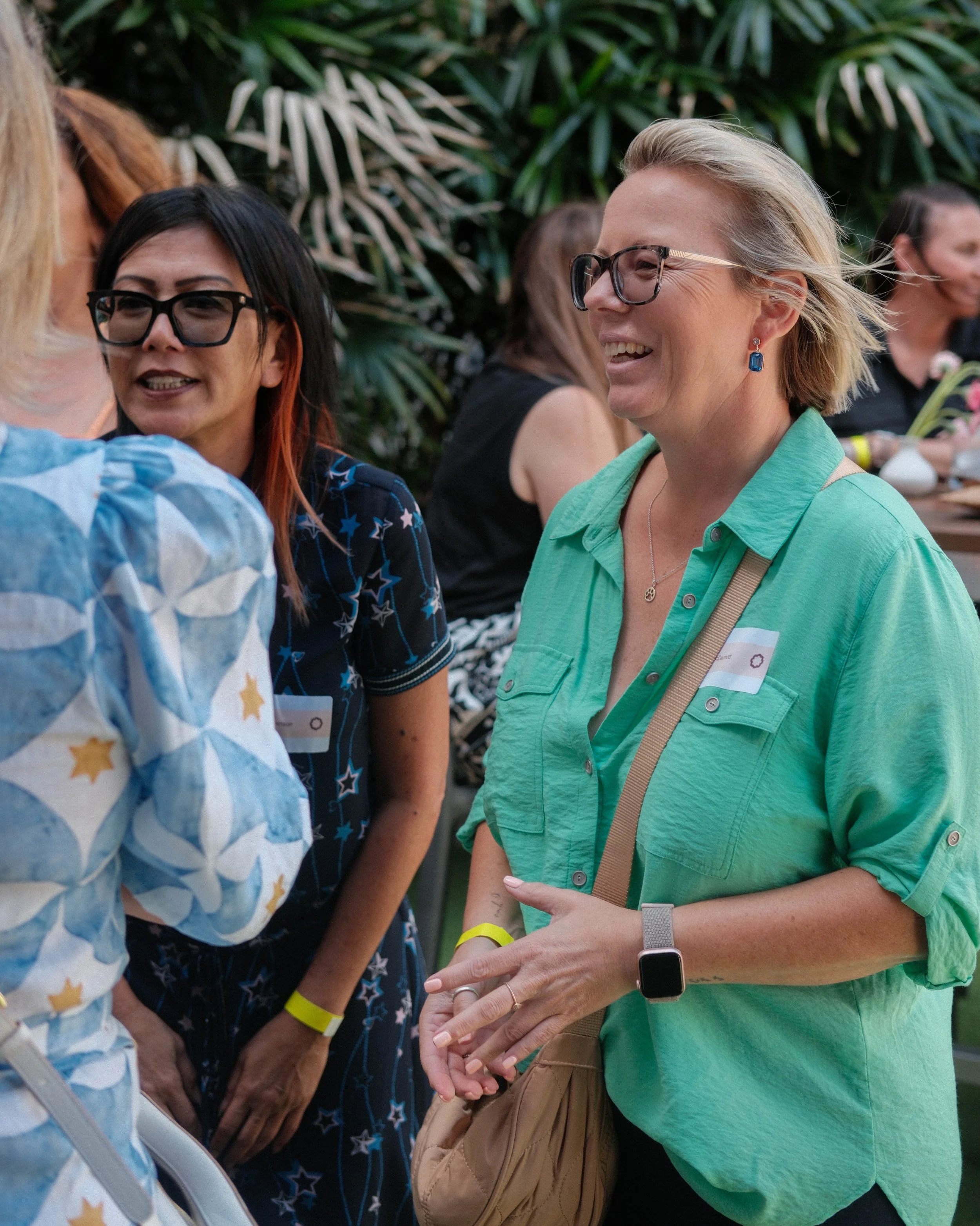 Group of women having a conversation outdoors, surrounded by green plants, smiling and wearing casual clothing and glasses.