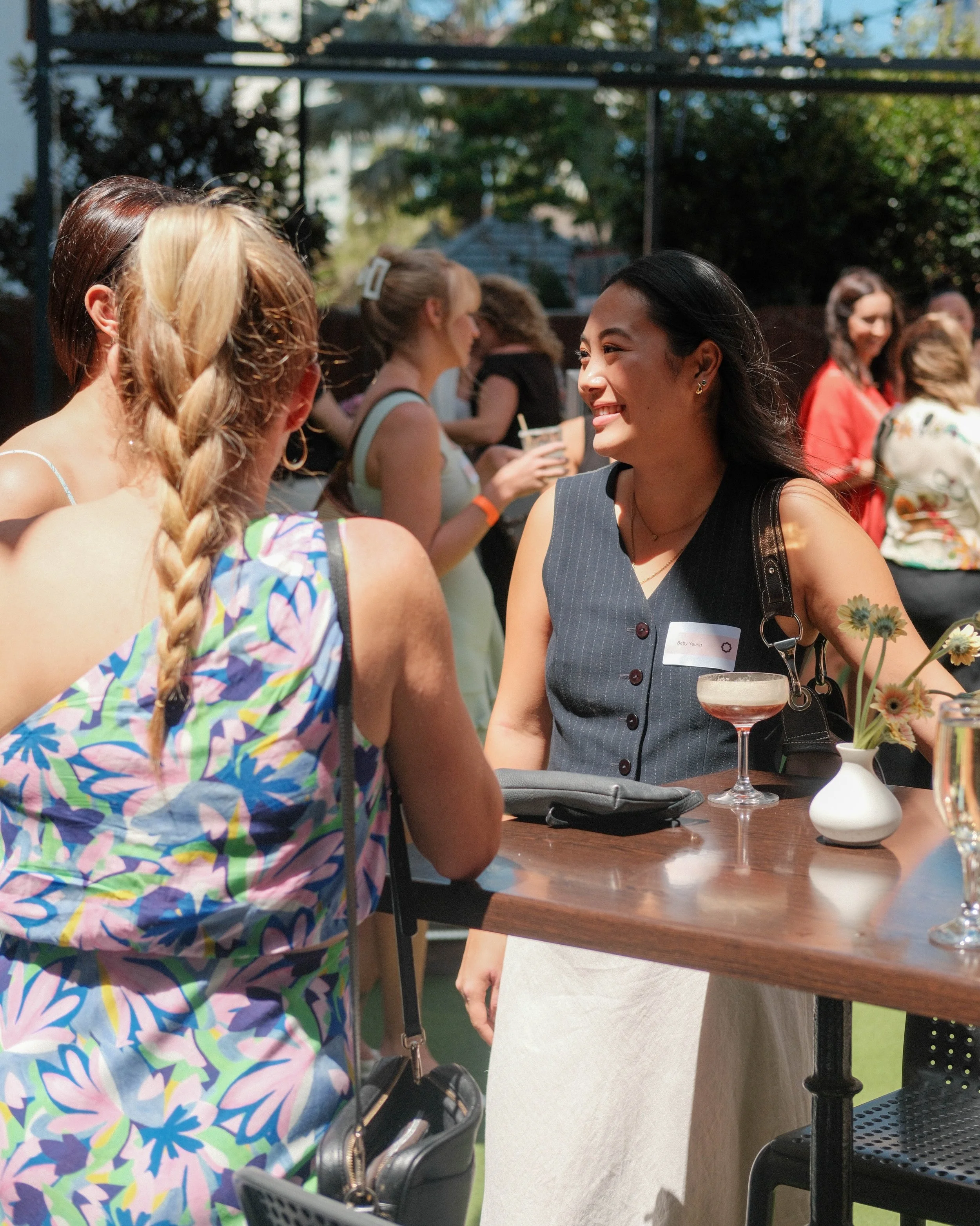 Women socializing outdoors during daytime at a gathering, smiling and engaging in conversation, with drinks on the table and a vase of flowers.