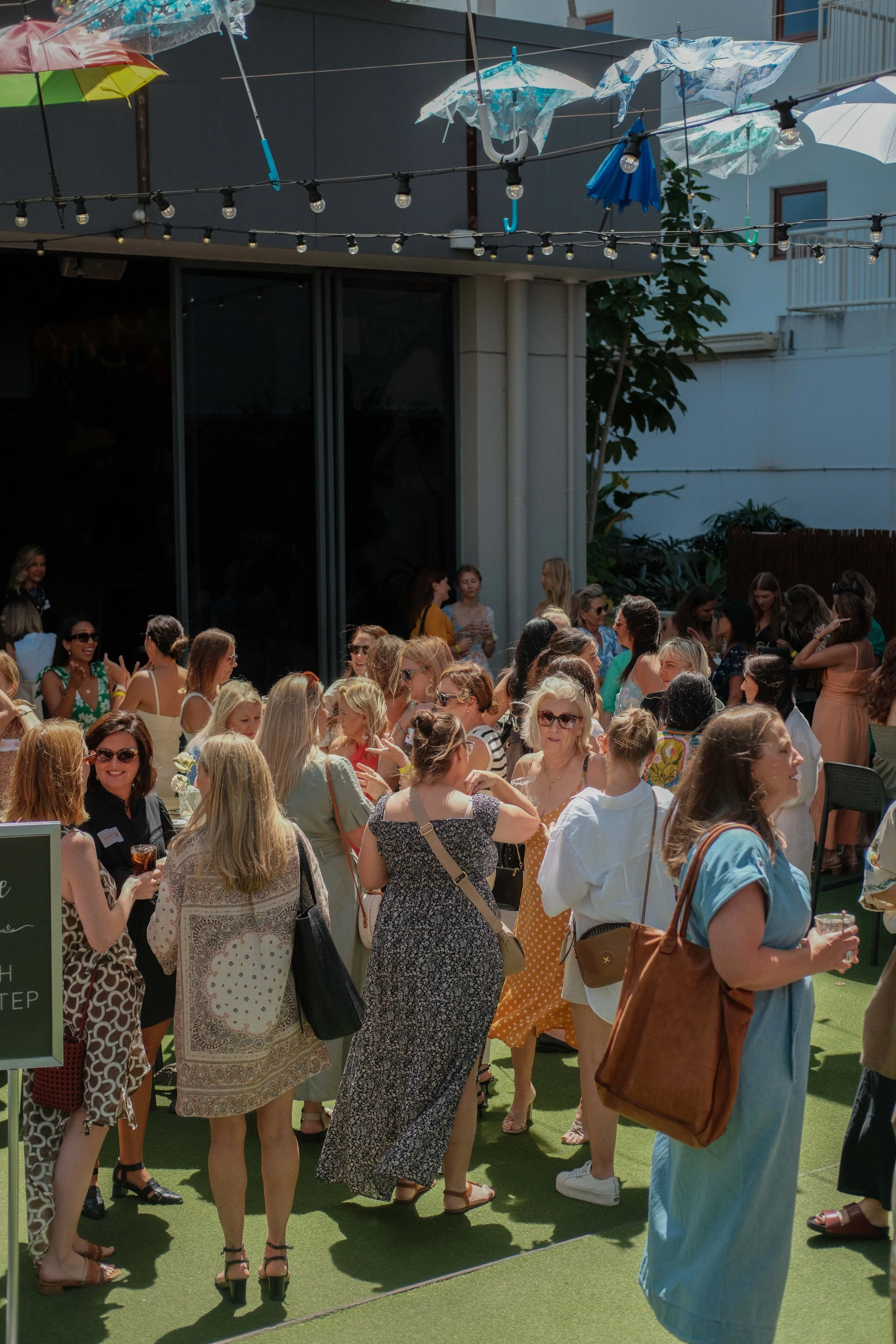 A group of women gathered outdoors on a sunny day with some holding drinks, socializing at an event with umbrellas hanging overhead and a building in the background.
