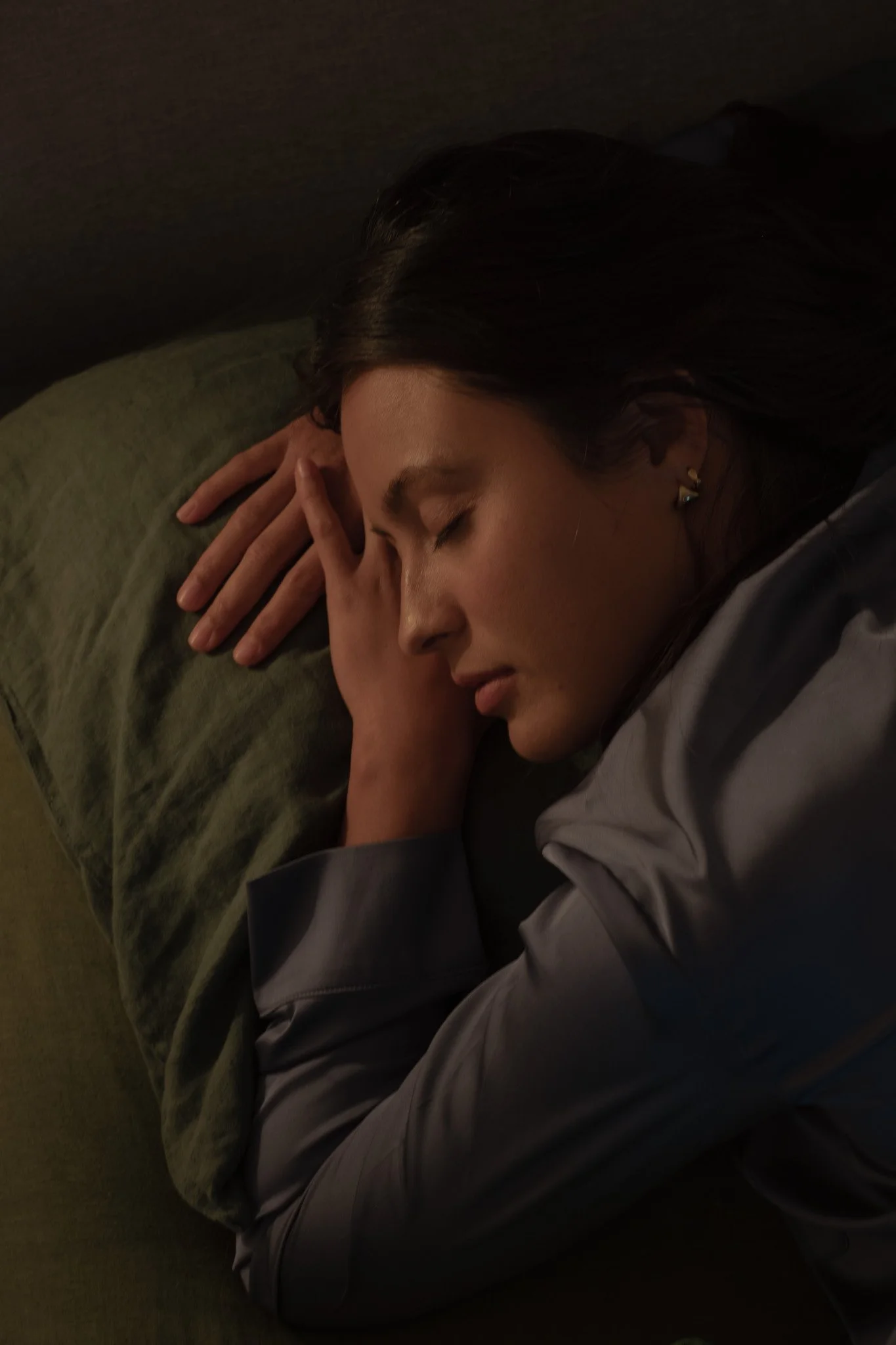 A woman is sleeping on a green pillow, lying on her side with her hands resting near her face, in a dimly lit setting.