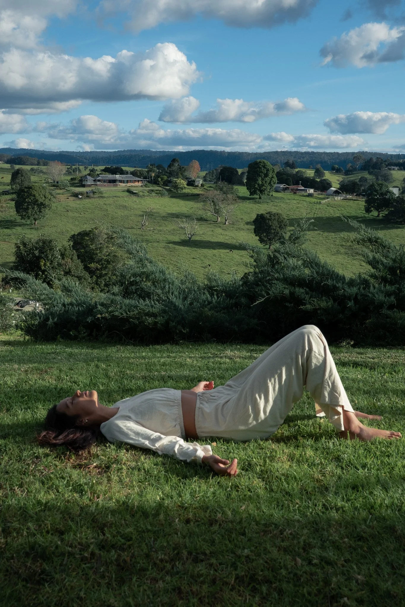 A woman with long dark hair sitting cross-legged on grass, her eyes closed, with one hand on her chest and the other on her stomach, in a peaceful outdoor setting with trees and mountains in the background.