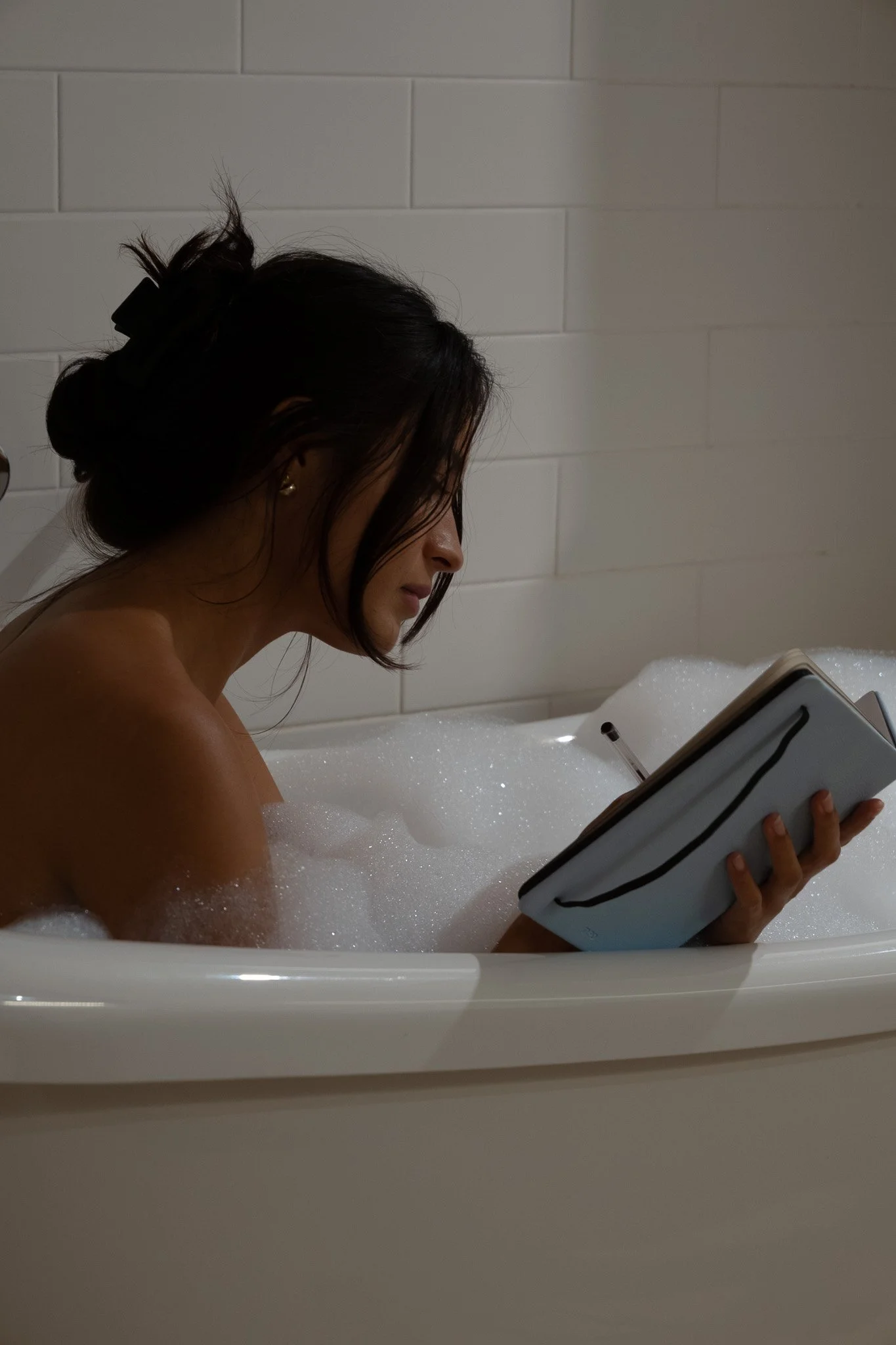 A woman with dark hair tied back, sitting in a bubble bath, reading a book.