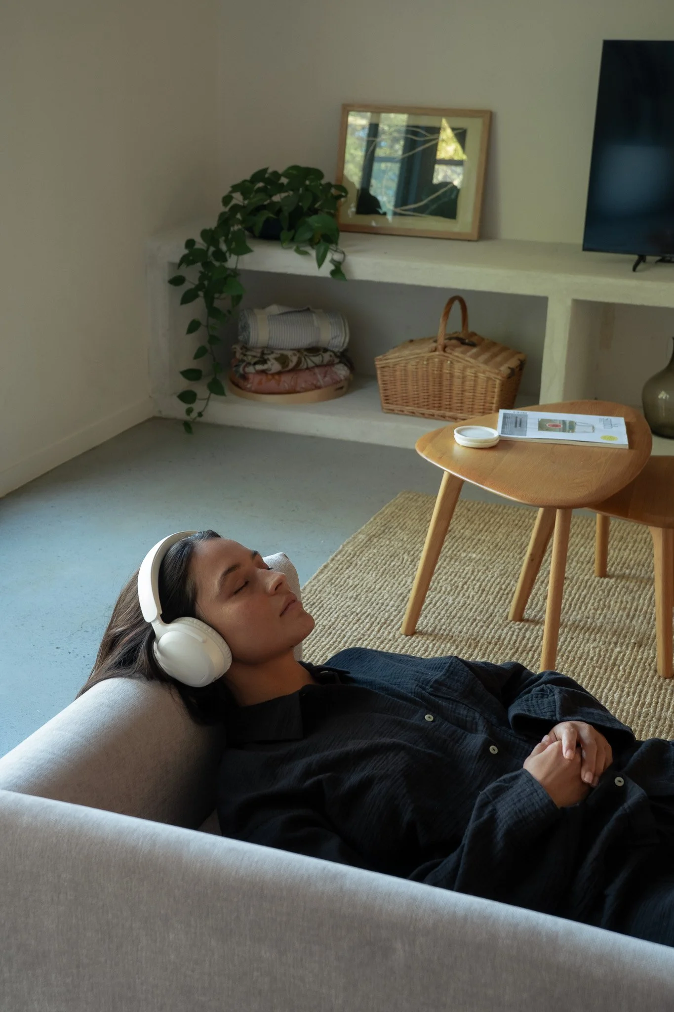 A woman with long dark hair relaxing on a light-colored couch, wearing white headphones with her eyes closed, in a cozy living room with a wooden side table, a beige rug, a shelf with a basket and folded blankets, a plant, and a mirror reflecting a window.