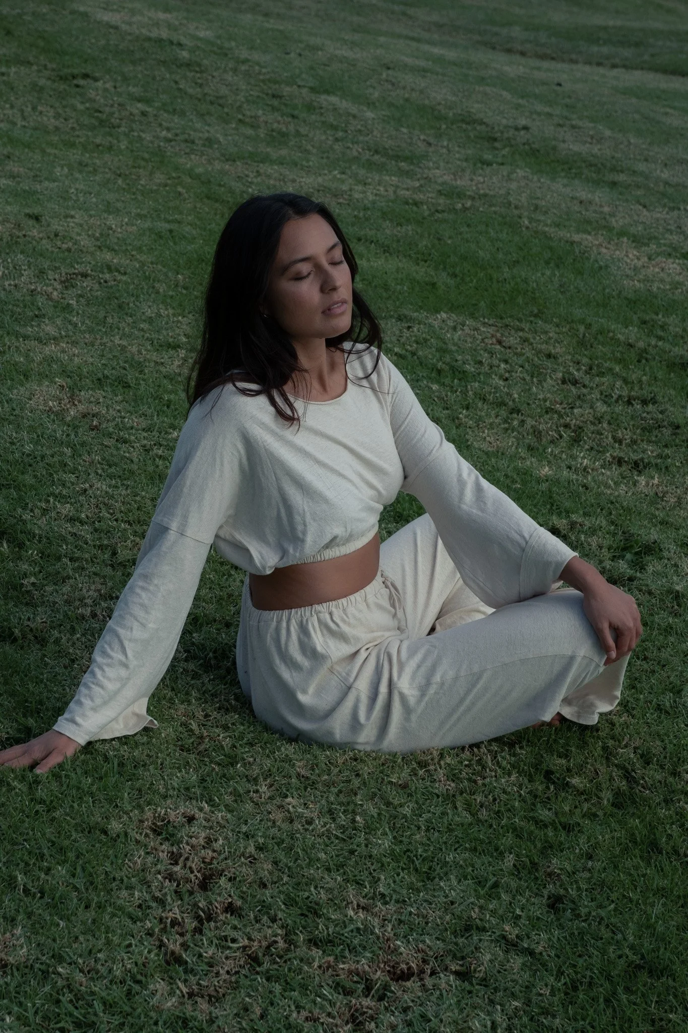 Woman sitting on grass with eyes closed, wearing a cream-colored long-sleeve top and matching loose pants.