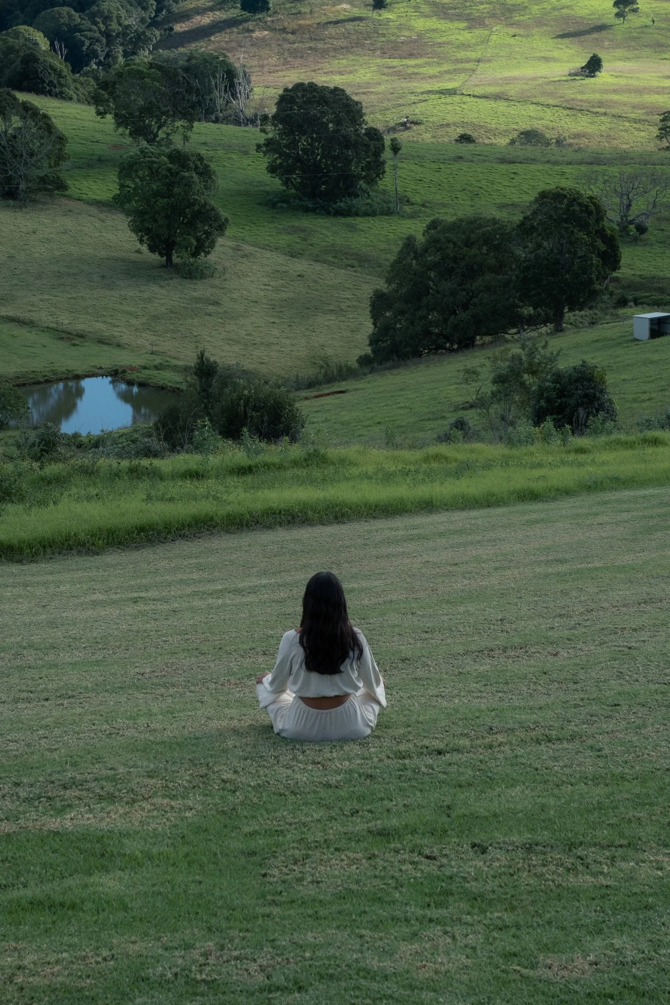 A woman with long dark hair is sitting cross-legged on the grass facing a rolling green hillside with trees, a pond, and a small barn in the distance.