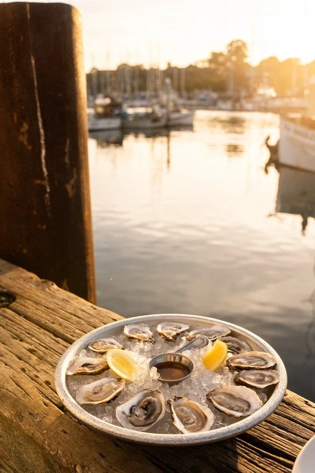 A metal platter of raw oysters on ice with lemon wedges, placed on a weathered wooden dock at a marina, with boats and water in the background during sunset
