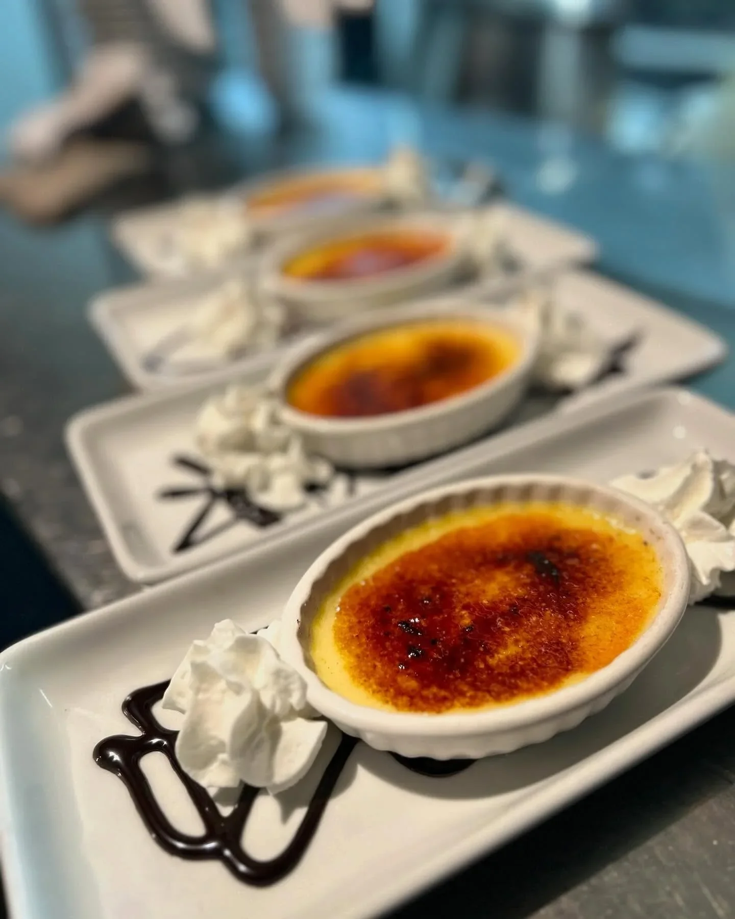 Three servings of crème brûlée on white rectangular plates decorated with whipped cream, chocolate syrup, and caramelized sugar on top.