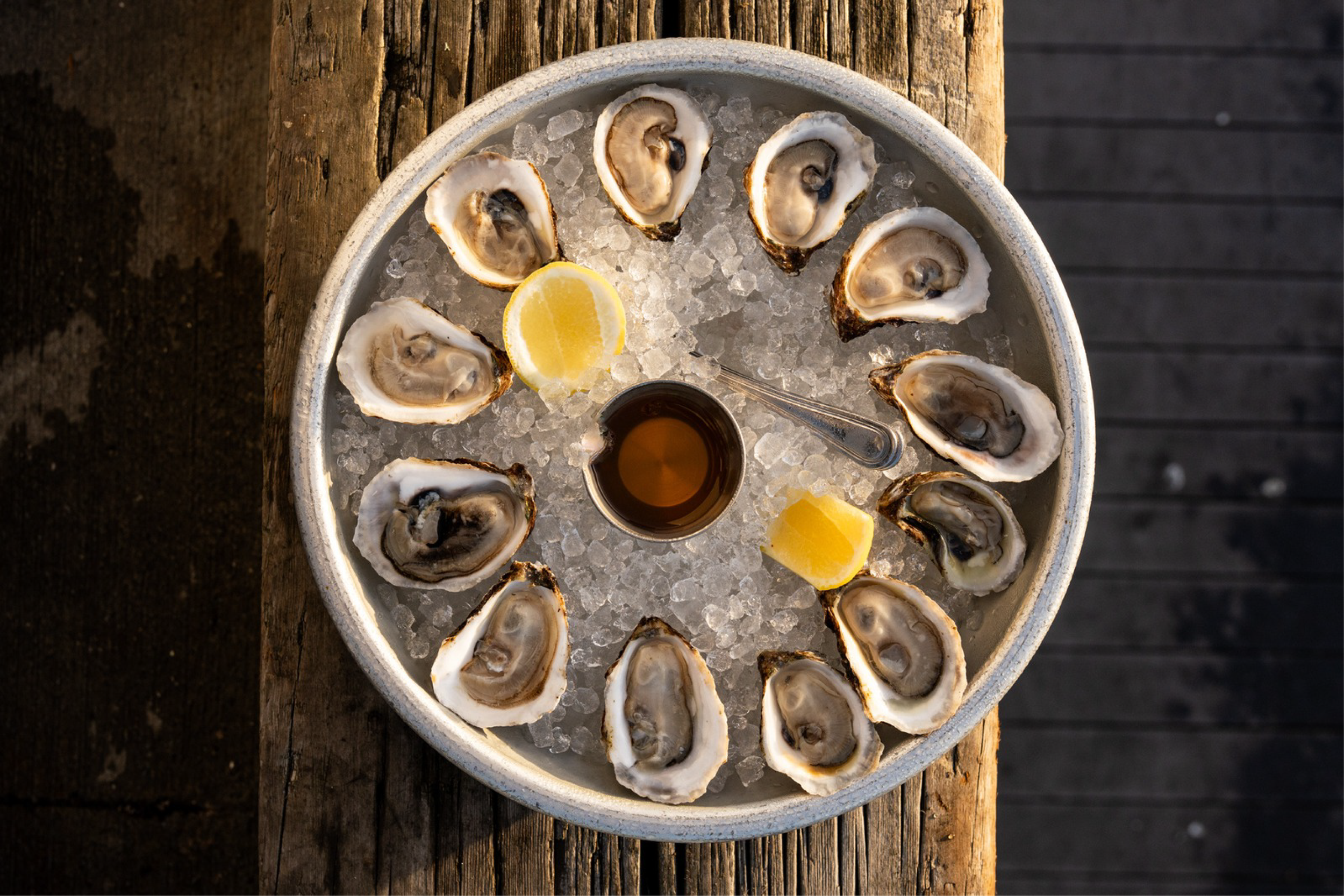 A round metal tray filled with ice, containing half shell oysters, lemon wedges, a small cup of mignonette, and a small cup of cocktail sauce, on a wooden surface.