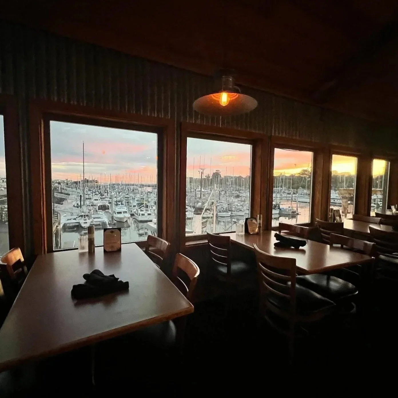 Interior of a restaurant with wooden tables and chairs, large windows showing a marina with boats and yachts at sunset.