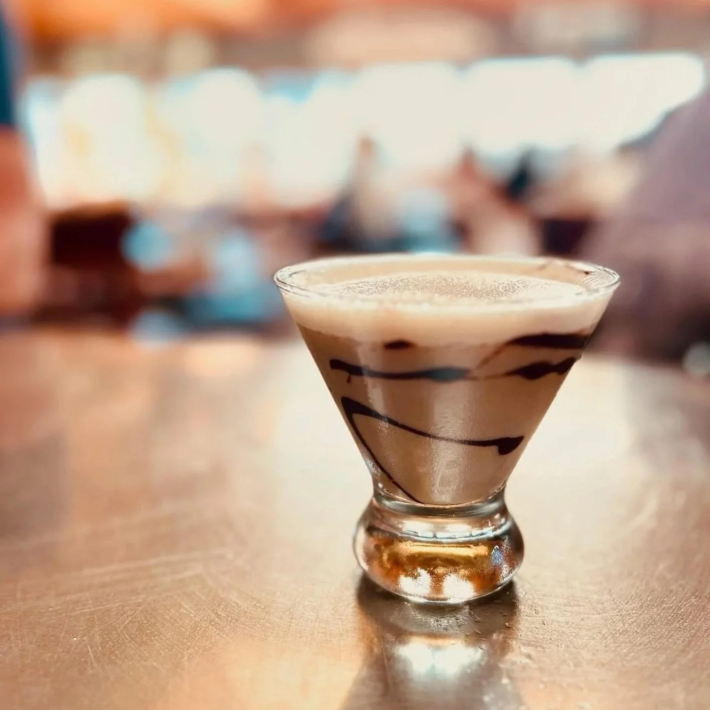 Close-up of a small glass with layered coffee drink, topped with foam and chocolate drizzle, on a wooden table with a blurred background.
