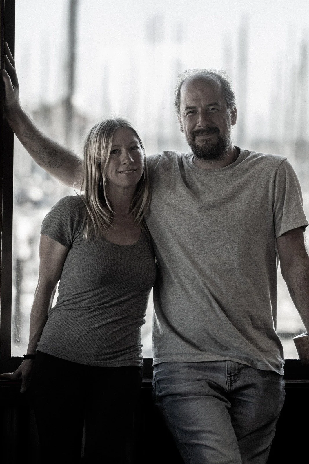 A woman and a man are standing together indoors near a large window, with the woman leaning slightly toward the man, who has his arm around her shoulders. They are smiling and appear relaxed.