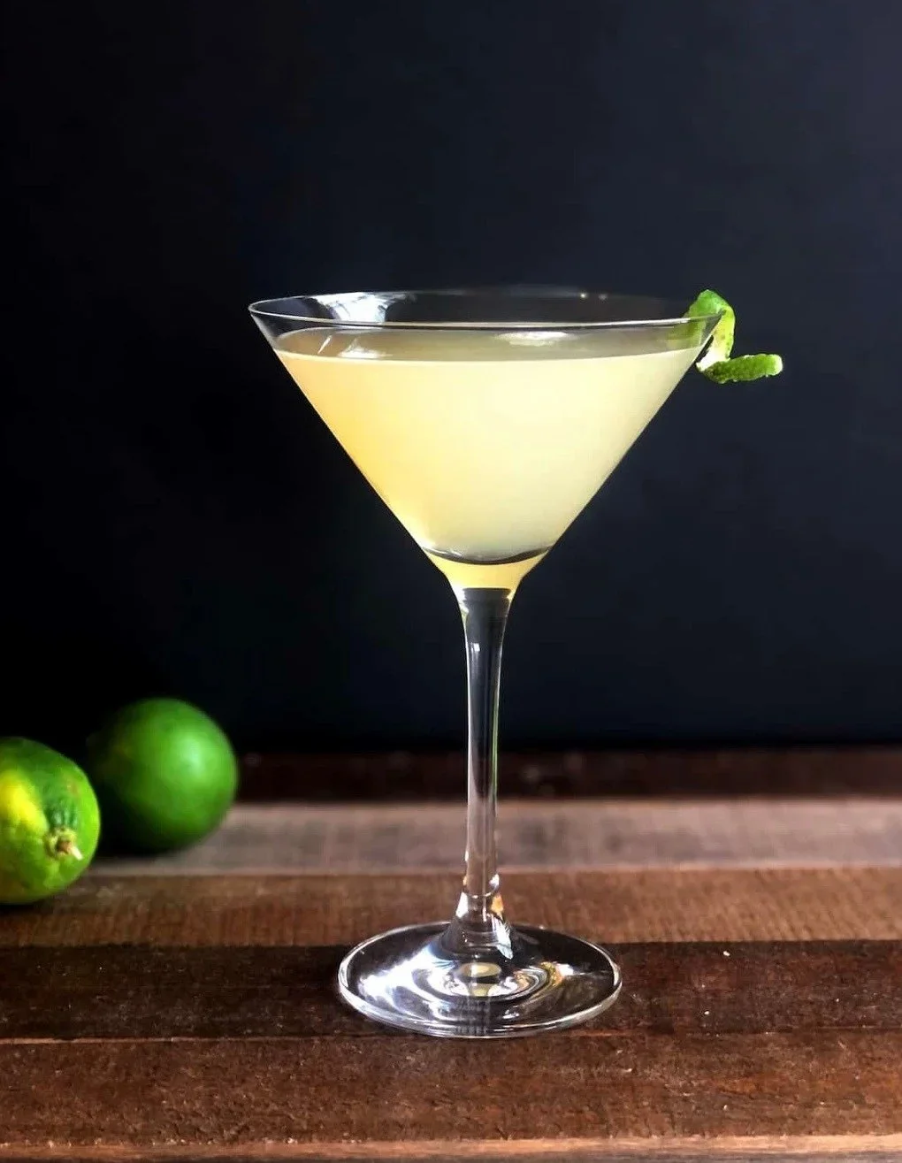 A frosty yellow cocktail in a martini glass garnished with a lime twist, with two limes on a wooden bar and dark background.
