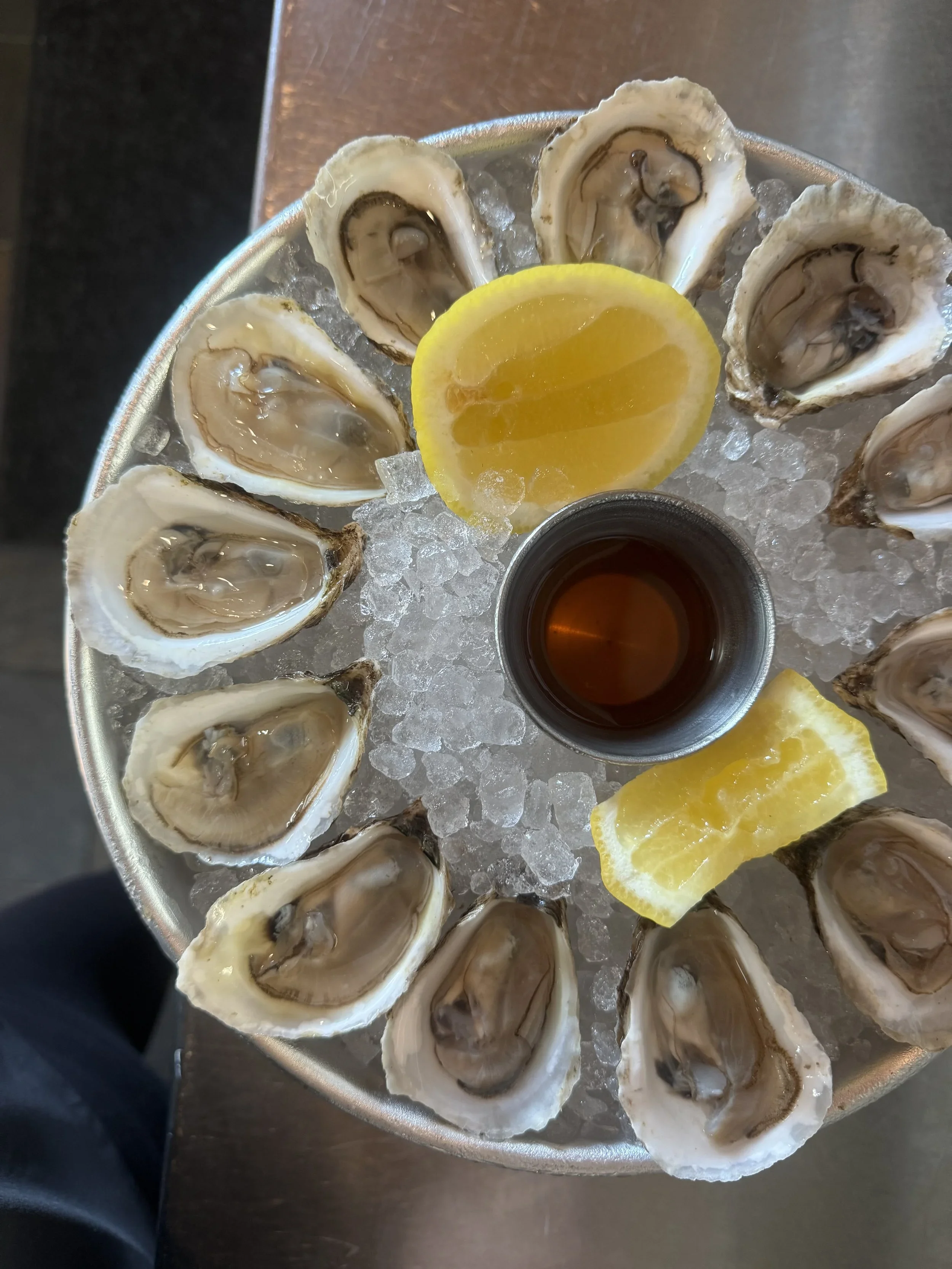 A round metal tray with ice, featuring 12 oysters on the half shell arranged in a circle, with lemon wedges, a small container of sauce, and a lemon slice in the center.