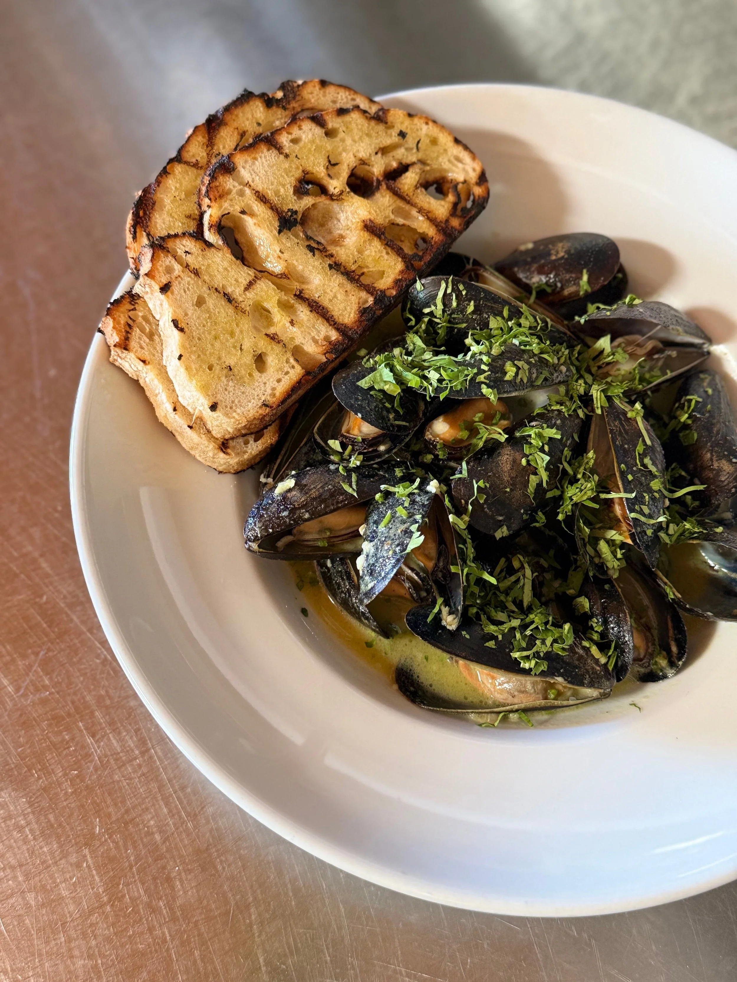 Sunset at a marina with boats docked on calm water, partly cloudy sky with orange and yellow hues, trees and buildings in the background.