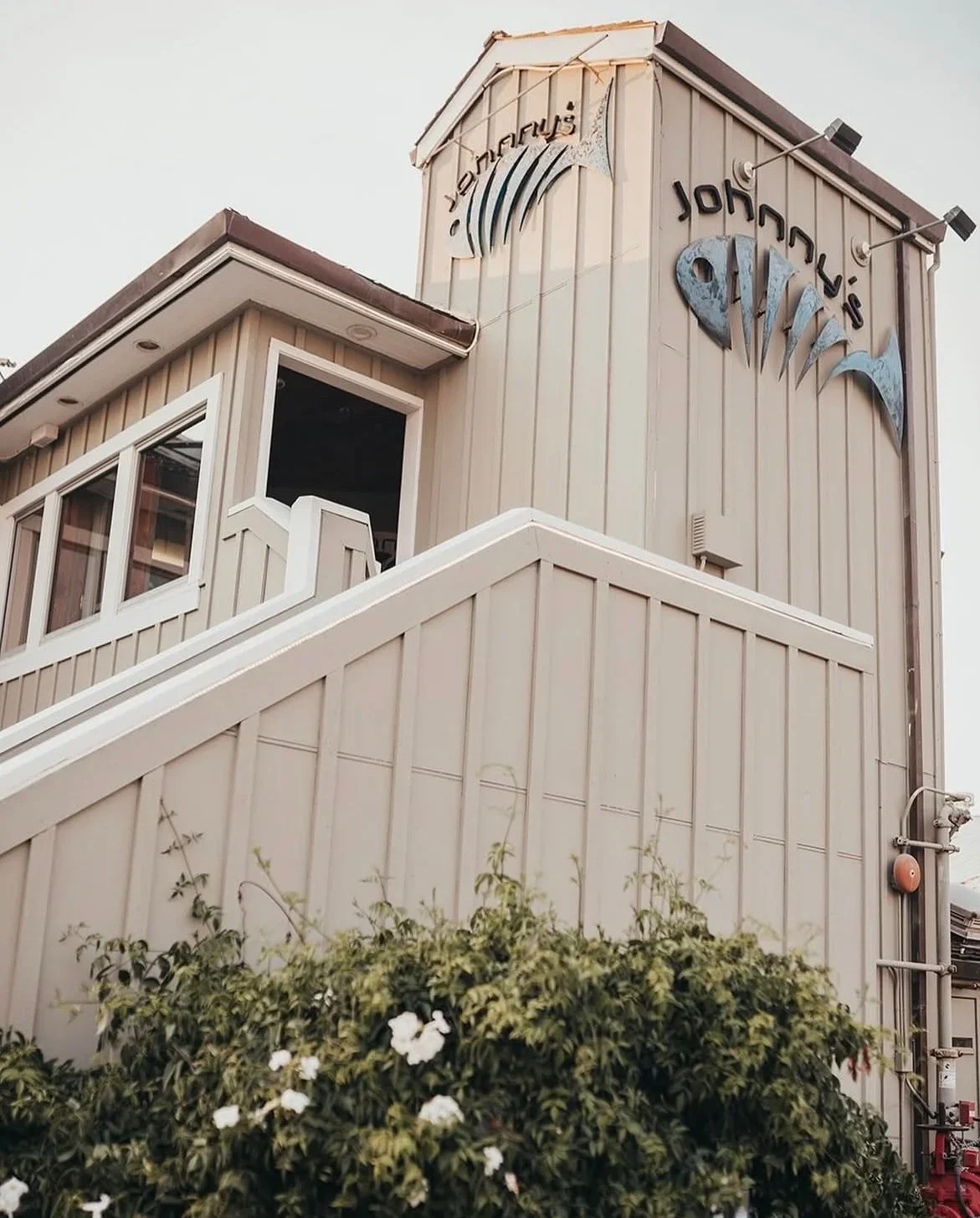 Exterior of the Johnny's Harborside Restaurant building with a sign that reads 'Johnny's' and a stylized fish logo and a rosebush with white blossoms at the entrance to the building's stairs.