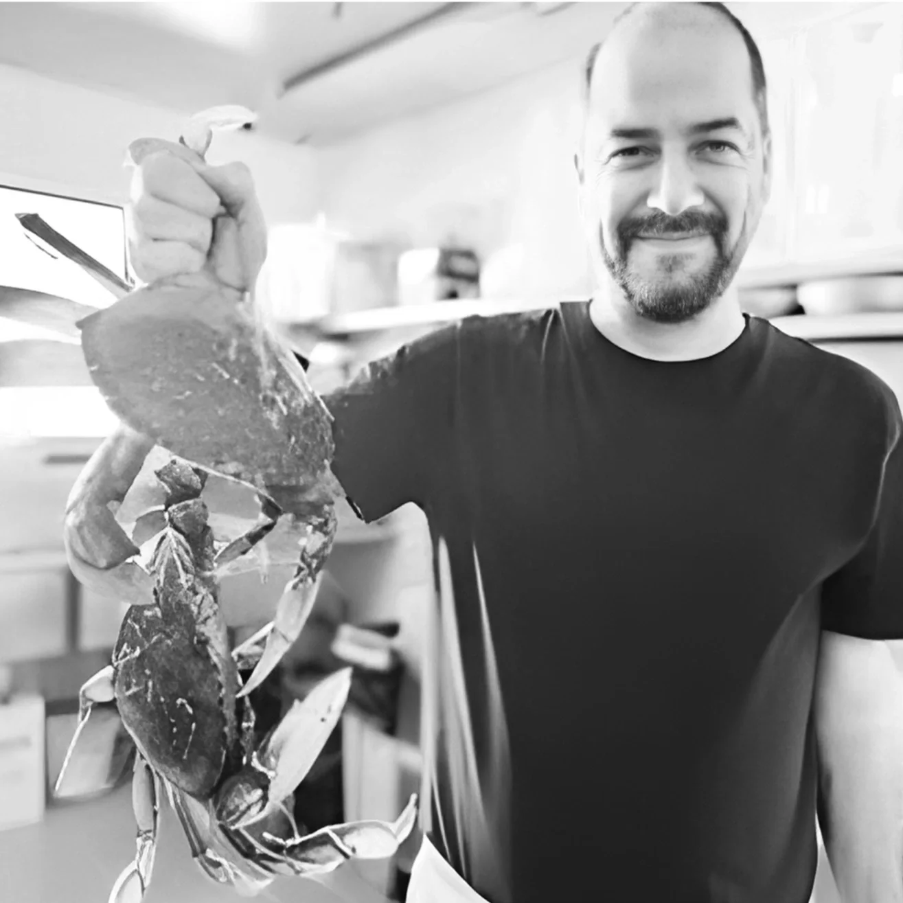 A man with a beard and short hair, wearing a black t-shirt and an apron, holding two large crabs in his hand, standing in the kitchen, and smiling at the camera.