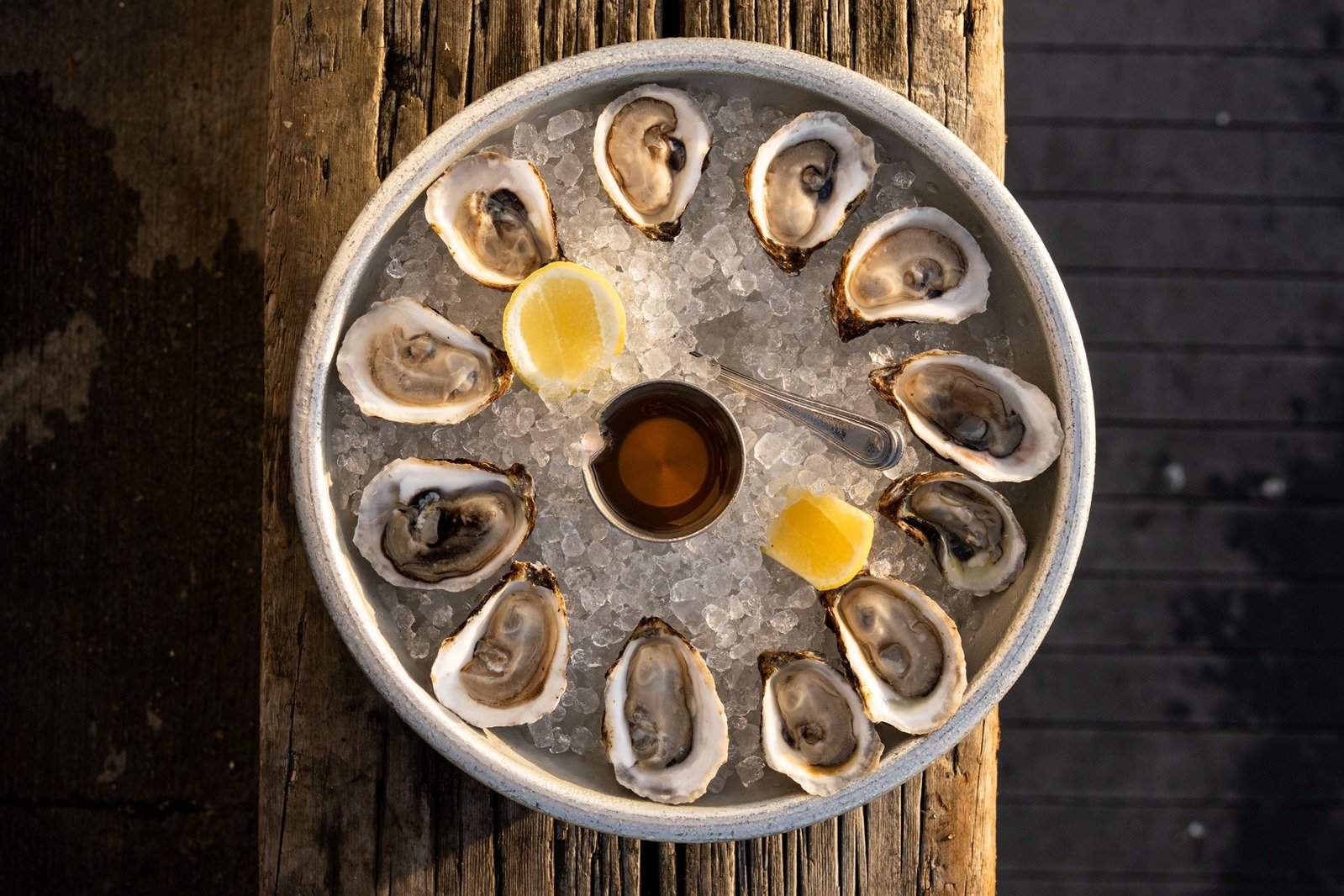 A round tray filled with ice, holding several oysters on the half shell, lemon wedges, a small cup of dipping sauce, and a small spoon.