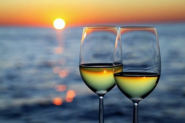 Two wine glasses filled with white wine on a table by the water, with a sunset in the background.