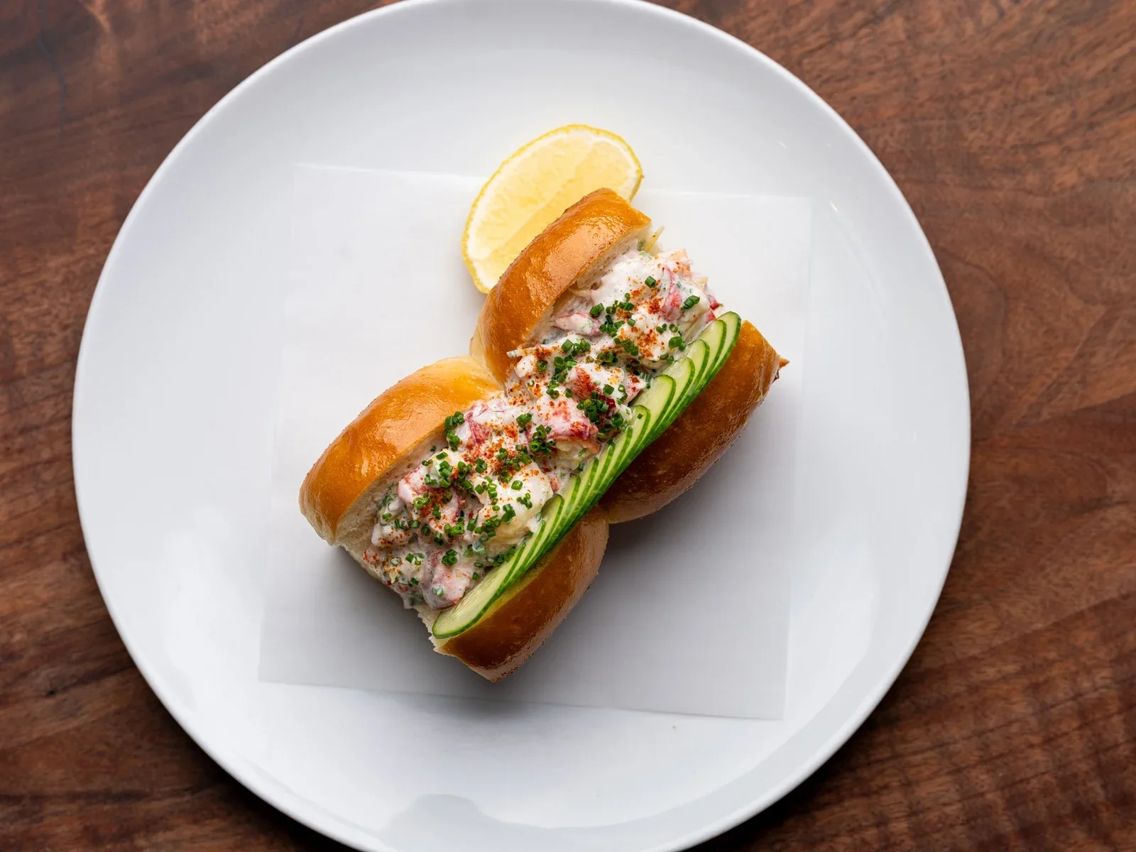 Lobster roll with chopped lobster meat, chives, and spices in a toasted bun, garnished with cucumber slices and a lemon wedge on a white plate.