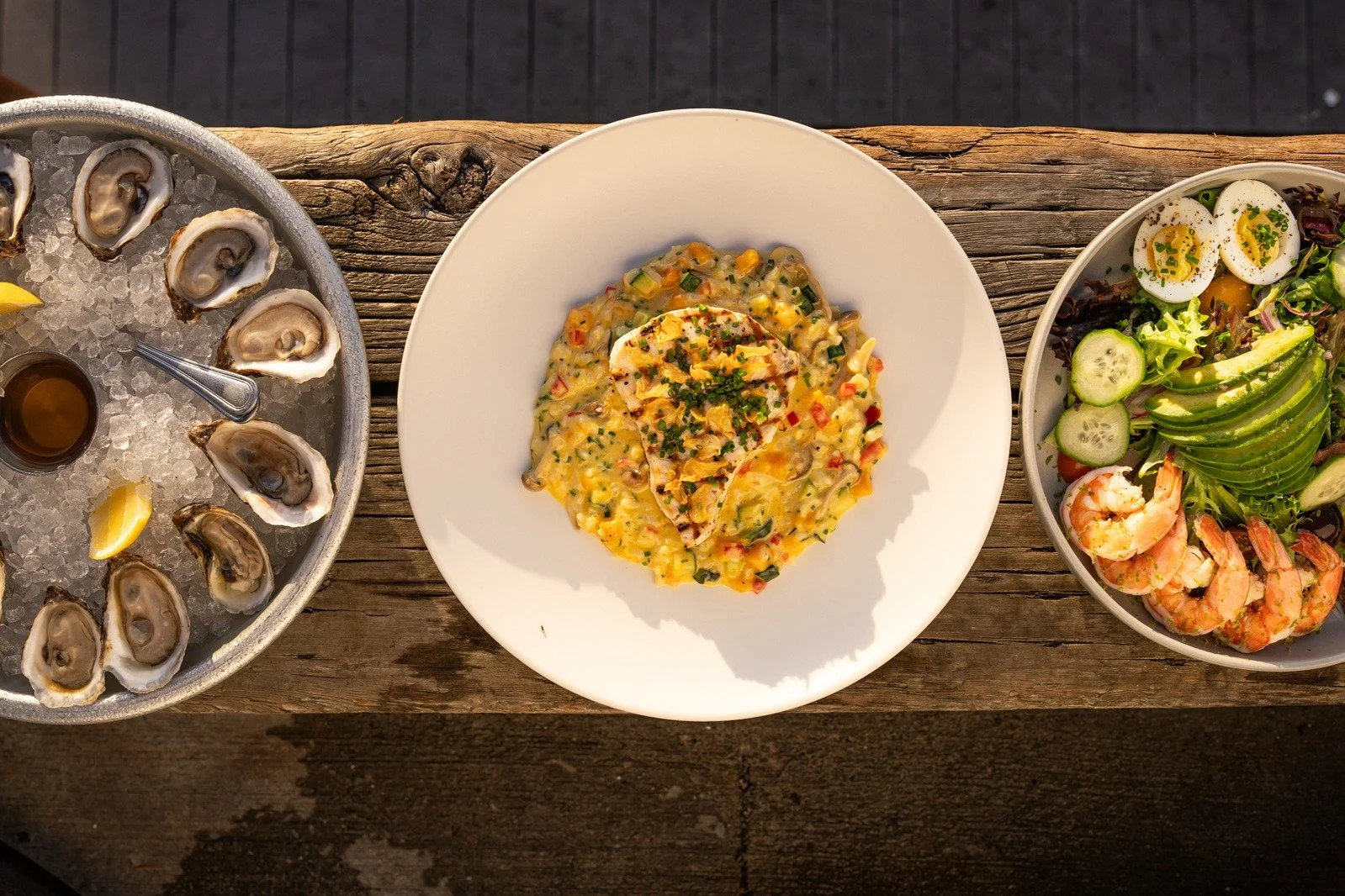 A top-down view of a rustic wooden table with three plates of food: on the left, a round silver tray of oysters on ice with lemon wedges and a small container of sauce; in the middle, a white plate with a creamy seafood dish topped with chopped chive