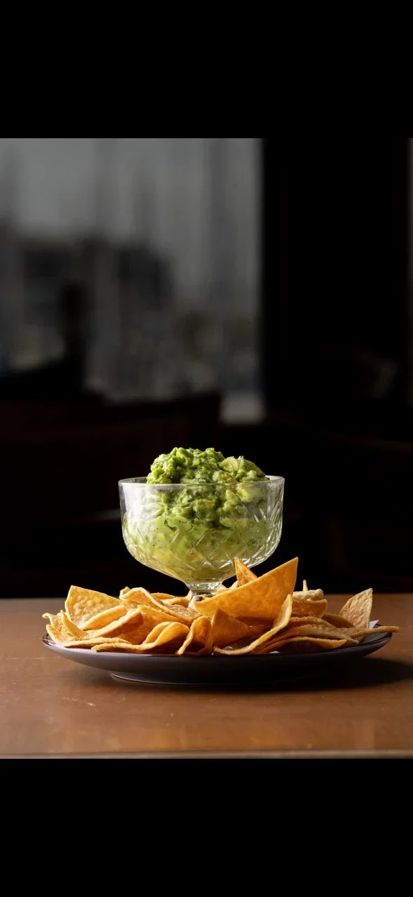 A glass bowl of guacamole on a plate of tortilla chips on a wooden table.