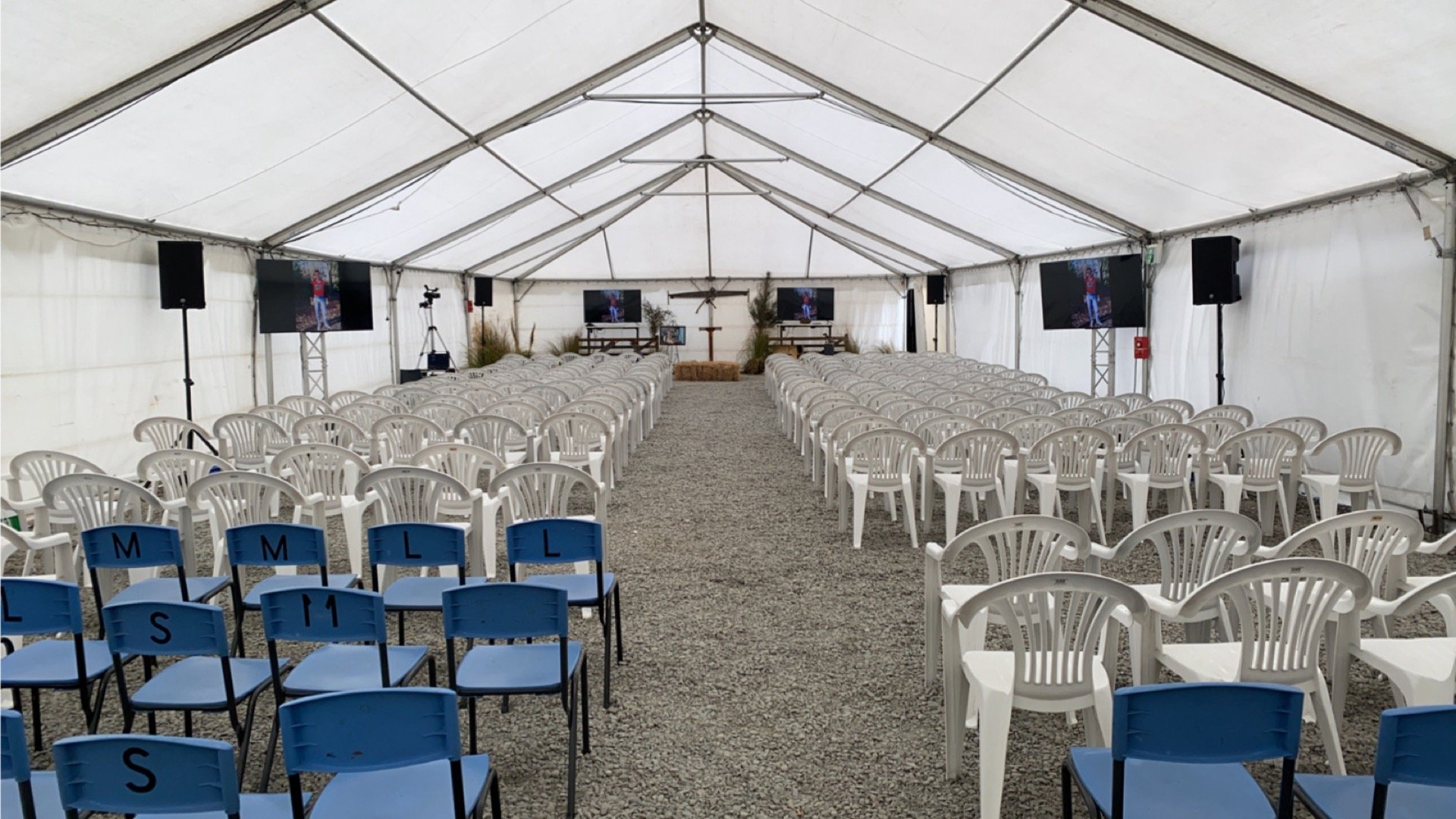 Empty tent setup with rows of white plastic chairs facing a stage, two screens, and a cross on the wall, suggesting a religious or memorial event.