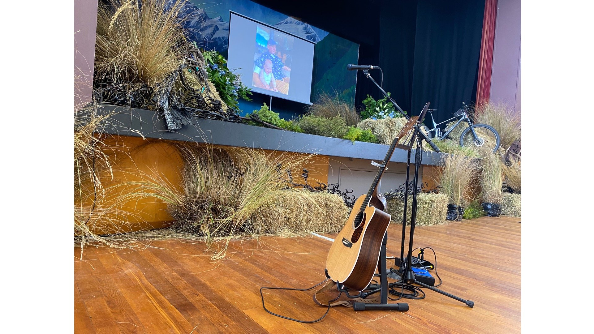 Stage setup with a microphone stand, acoustic guitar, and a projector screen displaying a family photo, surrounded by hay bales and dried grass decorations. A bicycle is partially visible on stage.
