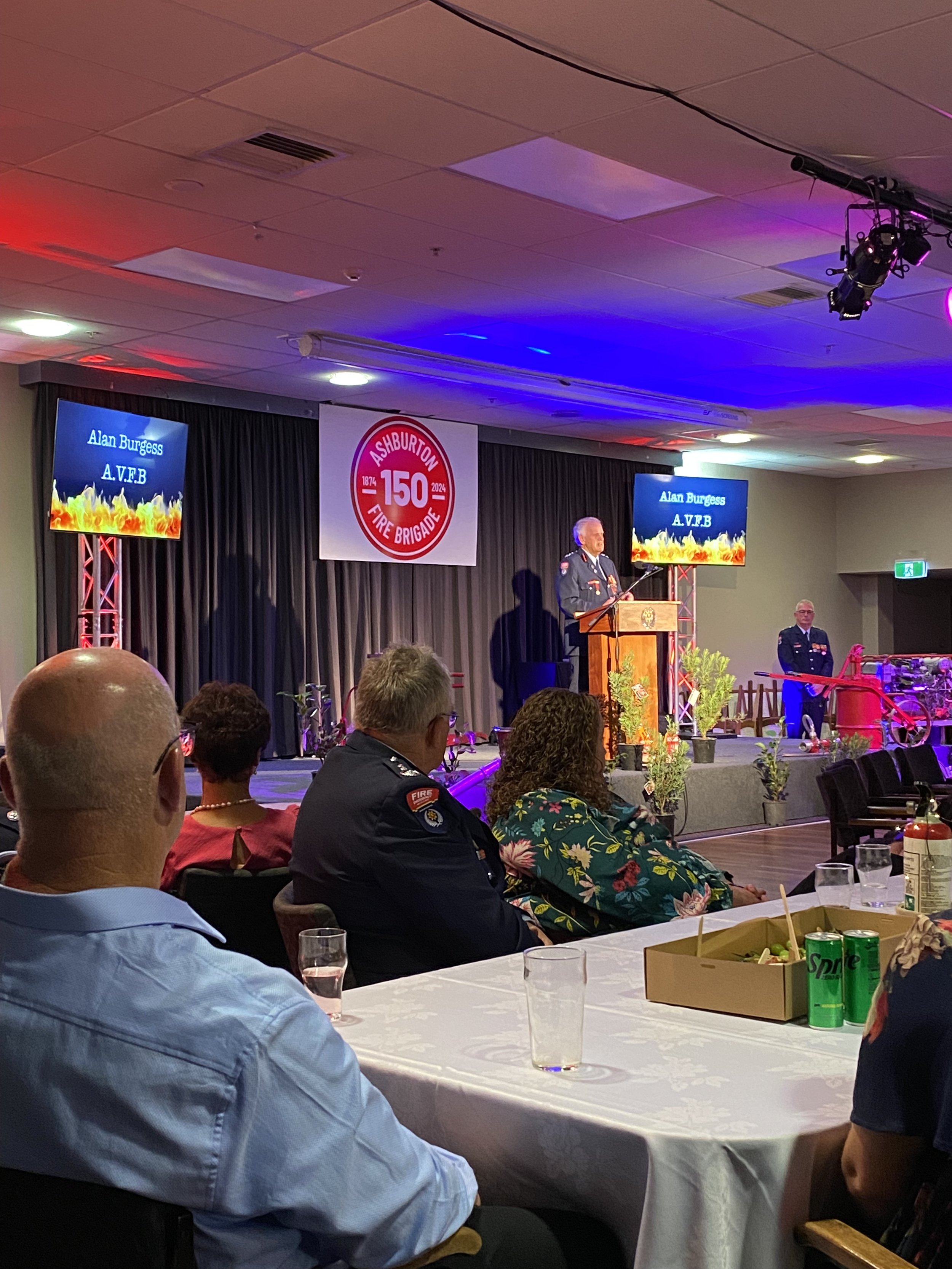 A person stands at a podium speaking at an event celebrating the 150th anniversary of the Ashburton Fire Brigade. The room has dim lighting with red and blue accents. Attendees are seated at tables, some in fire brigade uniforms, listening to the spe