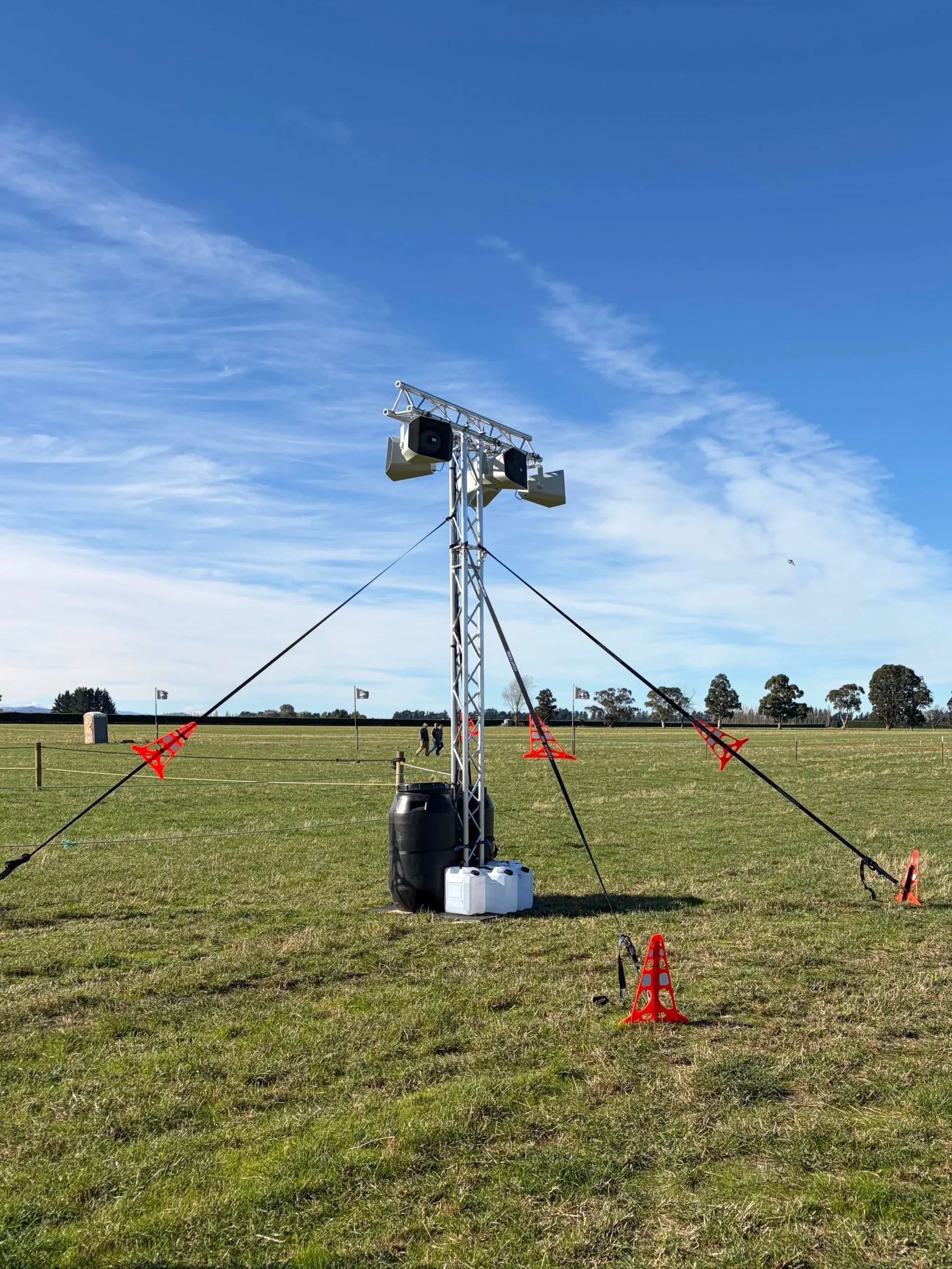 A temporary outdoor stage setup in a grassy field with a blue sky and scattered clouds. The stage includes a metal truss with mounted speakers and lighting equipment, supported by cords and orange safety cones.
