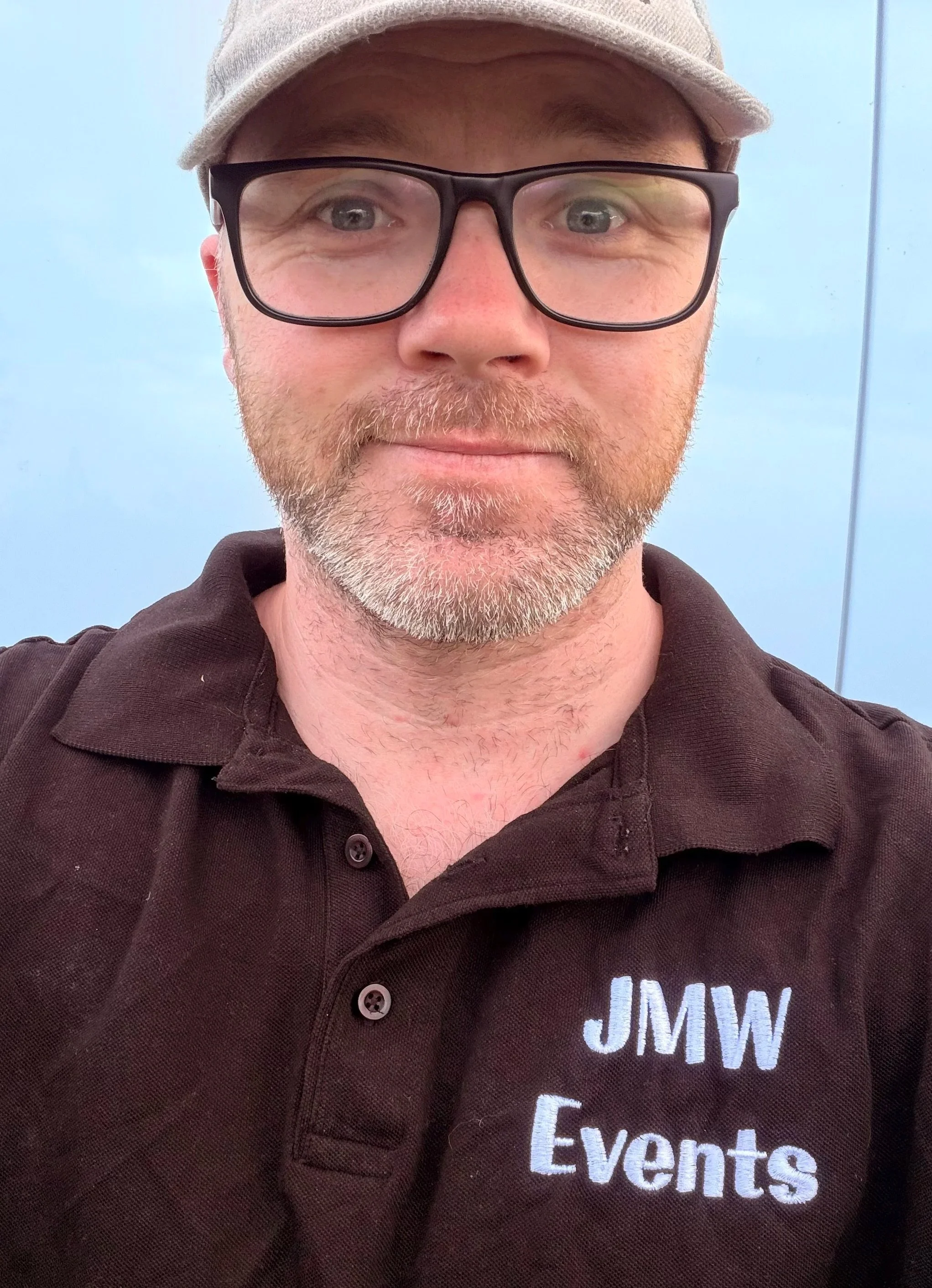 A man wearing glasses, a beige cap, and a brown shirt with 'JMW Events' embroidered on it, smiling against a light blue background.