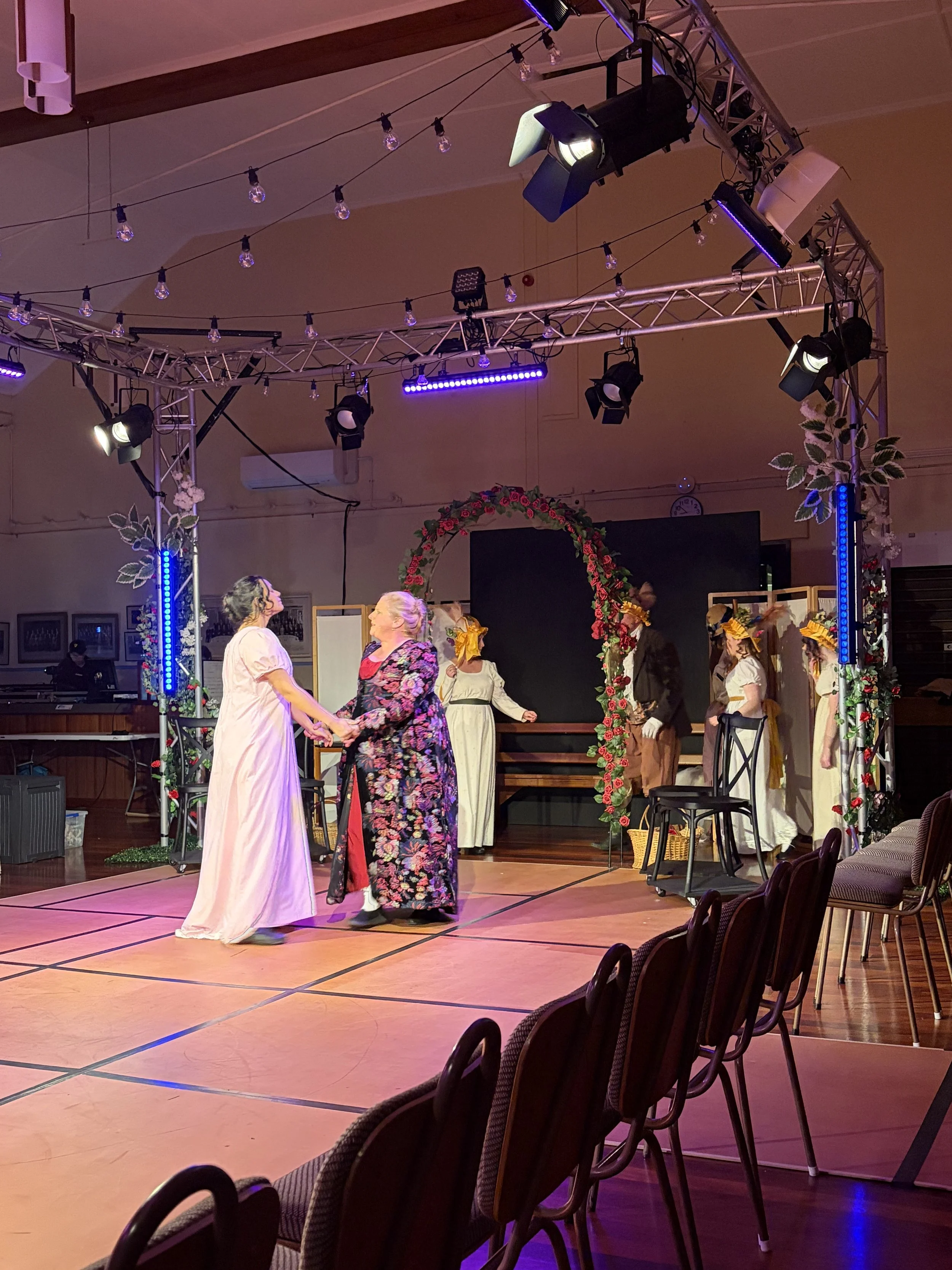 People on stage performing a dance in front of a decorative flower arch, with stage lights and hanging string lights overhead, in an indoor venue.