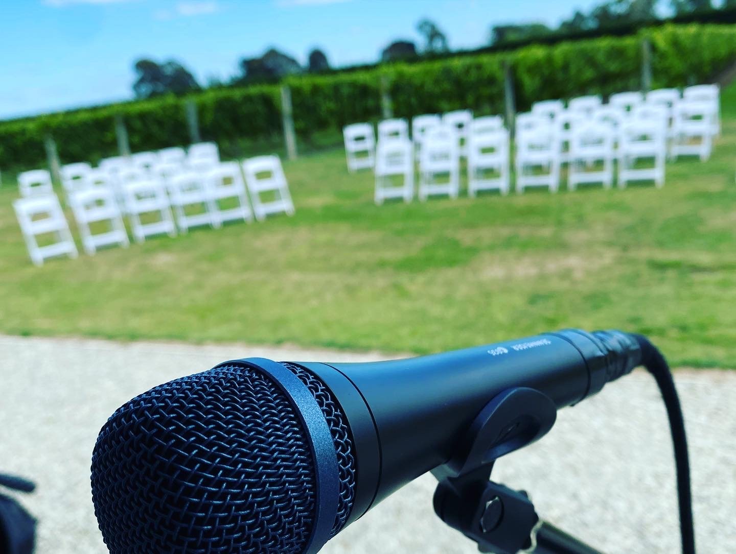 Microphone on a stand with white chairs set up outdoors on a grassy field, suggesting a public event or outdoor ceremony with vineyards in the background.