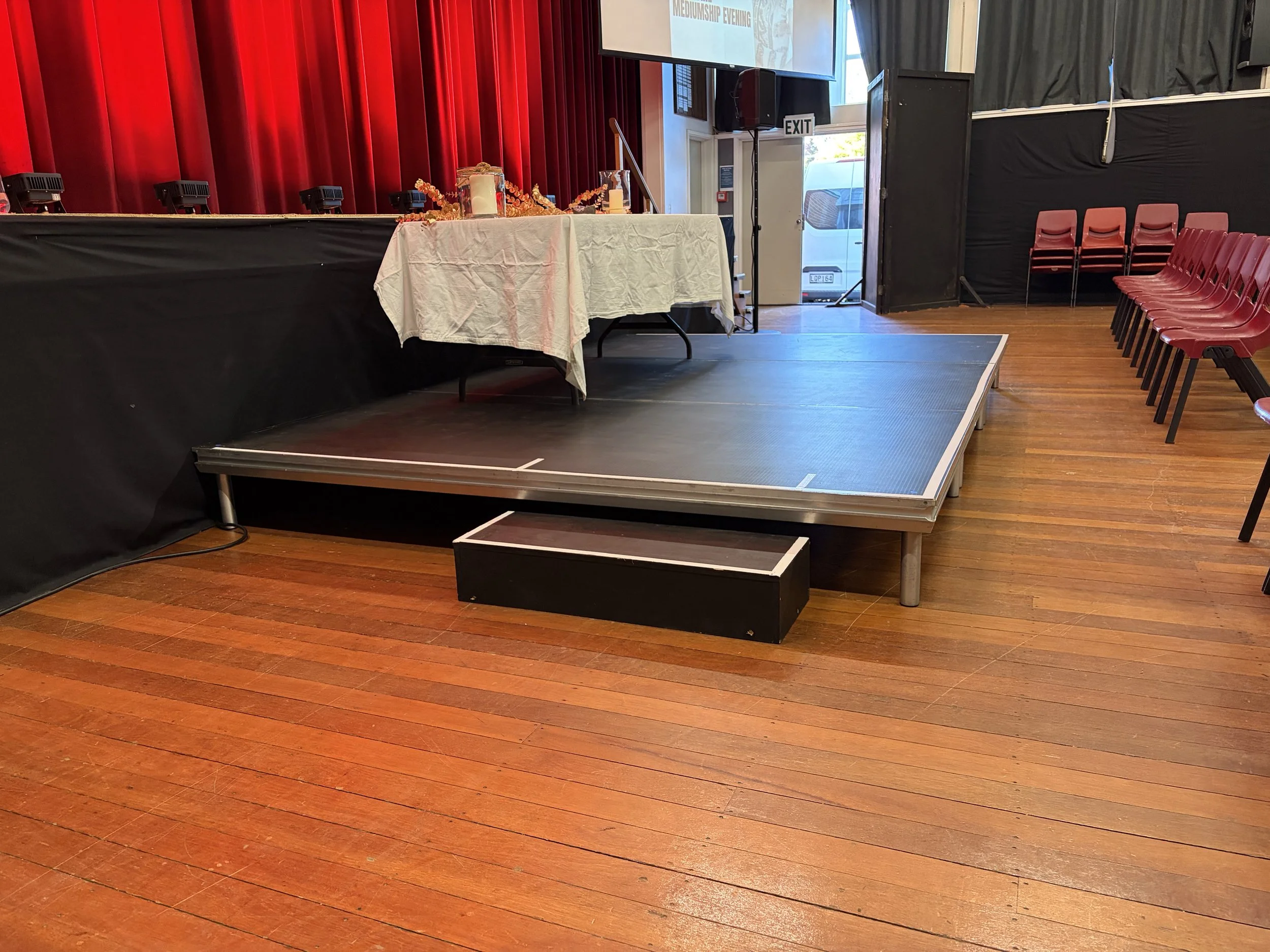 An empty auditorium with a small stage, red curtains, red chairs on the right, and a table with decorative items and candles near the stage. There is a large screen above the stage and an exit door behind the stage.