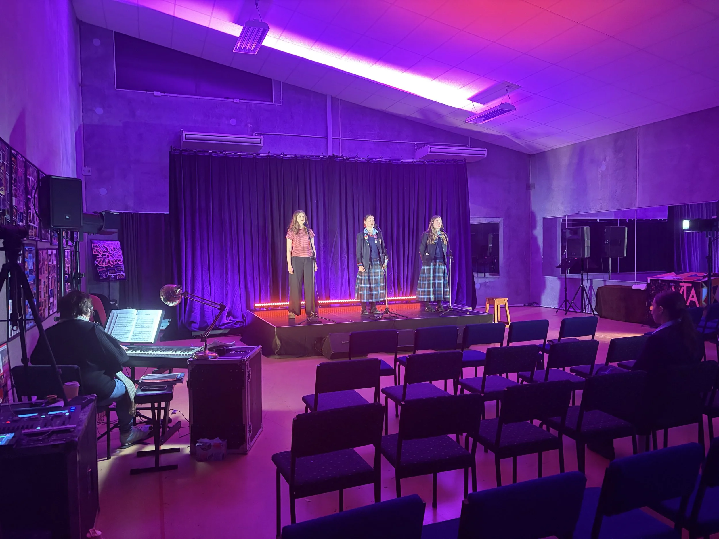 Three young women wearing plaid skirts and sweaters perform on a stage under purple lighting, with a black curtain backdrop in an indoor auditorium.