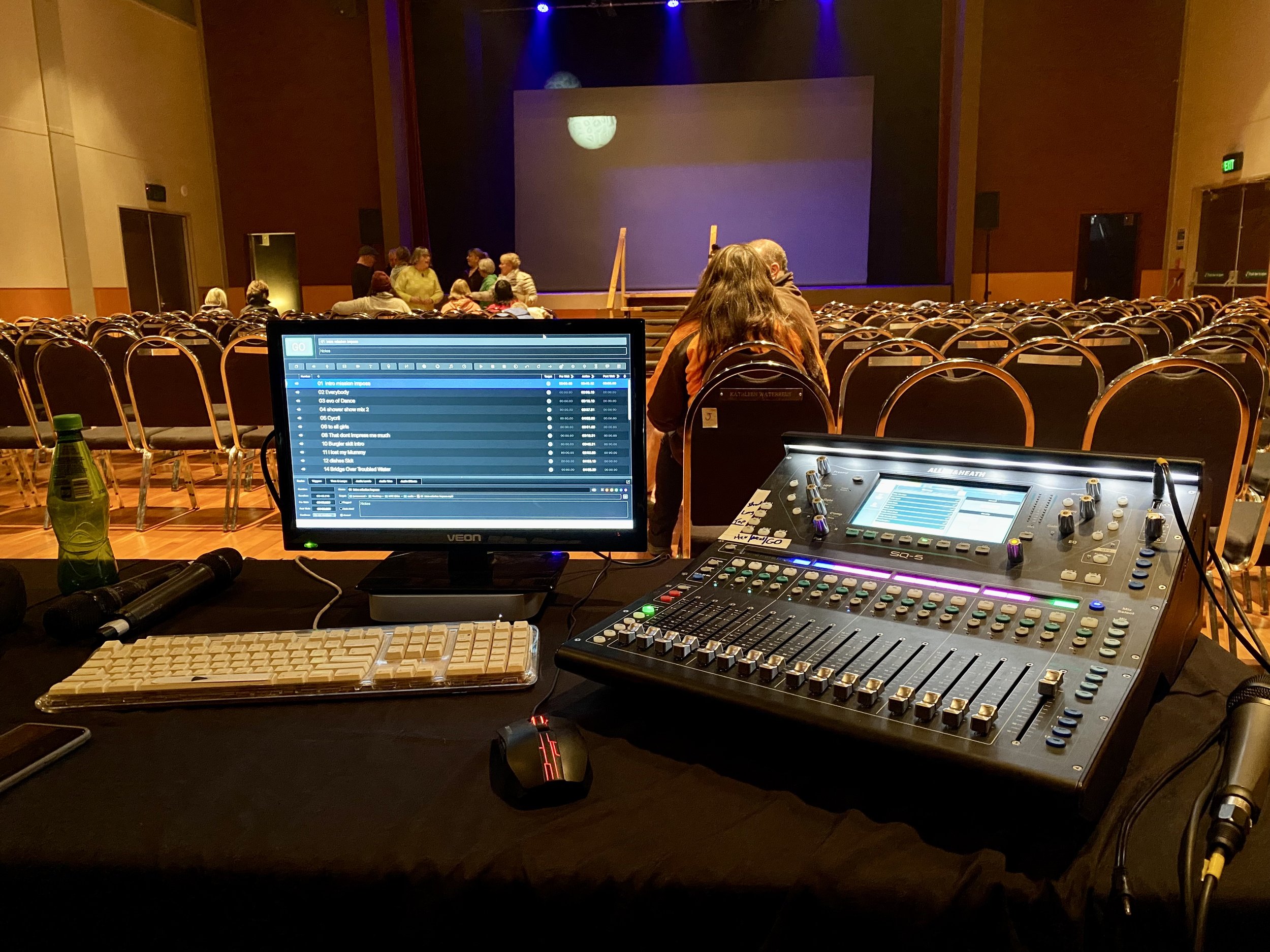 Audio mixing console and computer setup in a theater with seated audience and stage background.