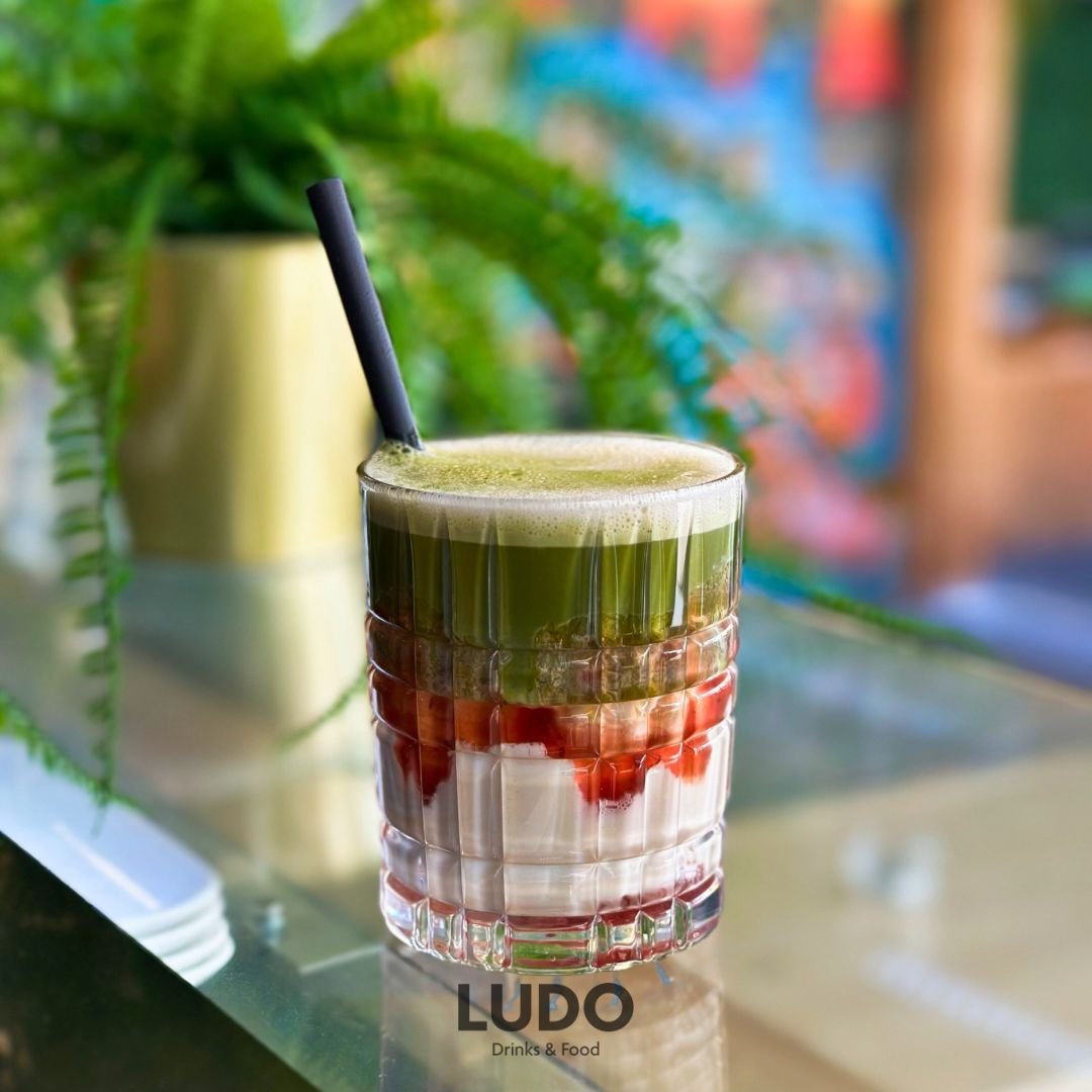 Ein mehrstöckiger Matcha mit grüner, roter und weißer Schichtung in einem Glas mit Strohhalm, im Hintergrund grüne Pflanzen und bunte Wand.