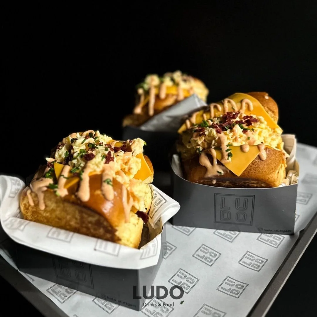 Drei Hot Dogs mit Kartoffelbrei, Käse, Speck und Saucen in schwarzen Papptellern auf einem LIDI Clear-Bapier mit LUDO-Logo, vor schwarzem Hintergrund.