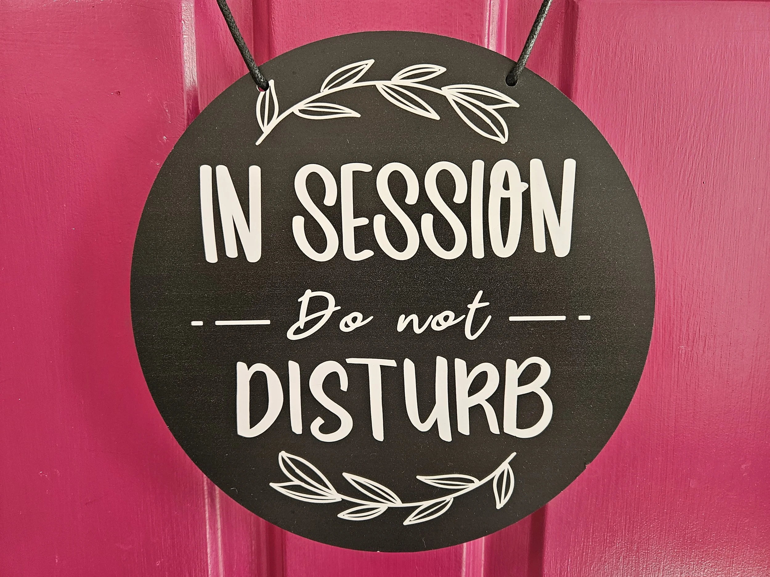 Black circular sign with white text that says 'In Session, Do not Disturb' and decorative white leaves at the top and bottom, hanging on a pink wall.