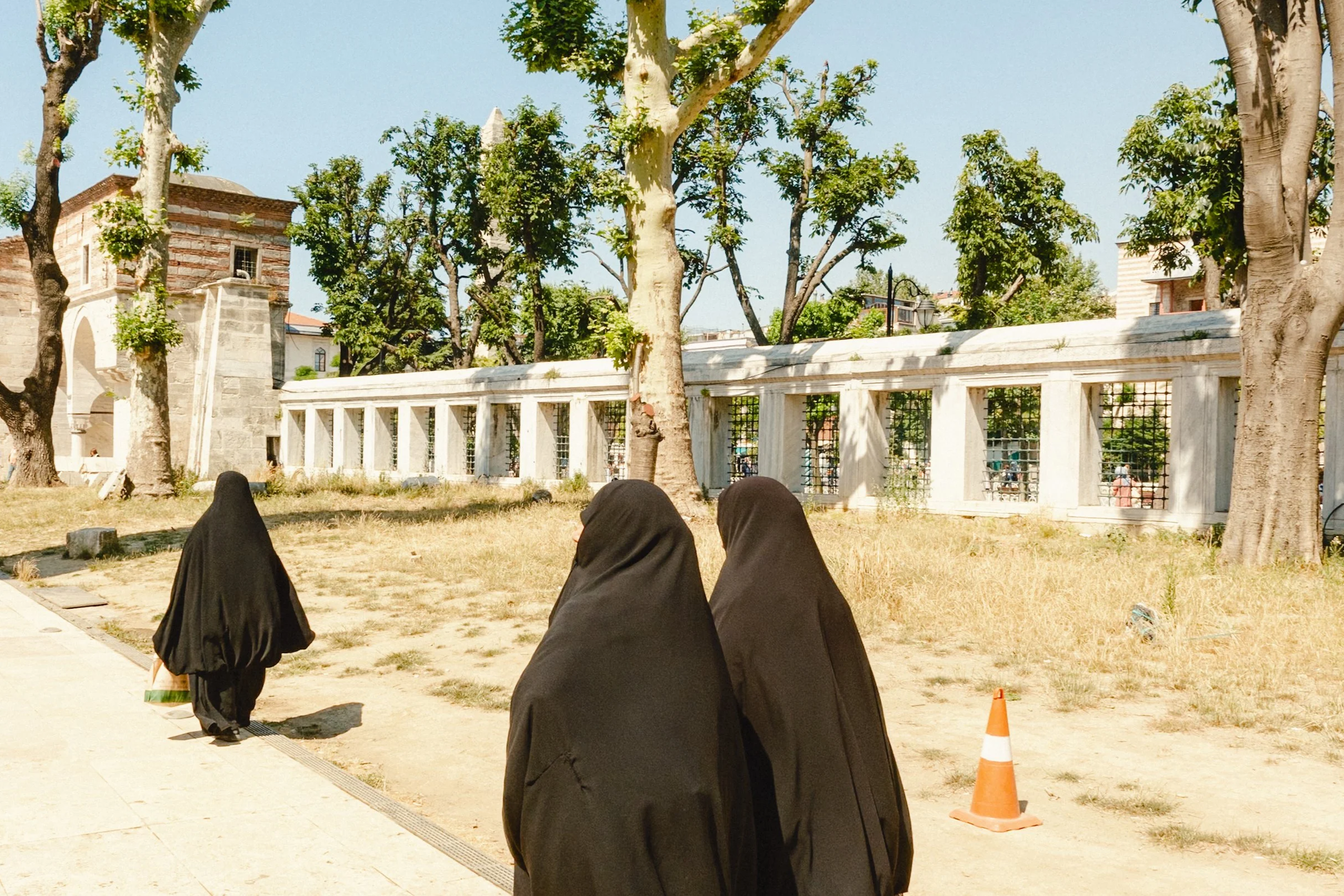 Women dressed in black walking near historic stone walls and trees in Istanbul, Turkey, under bright summer sunlight — candid Istanbul street photography showing cultural and everyday life.