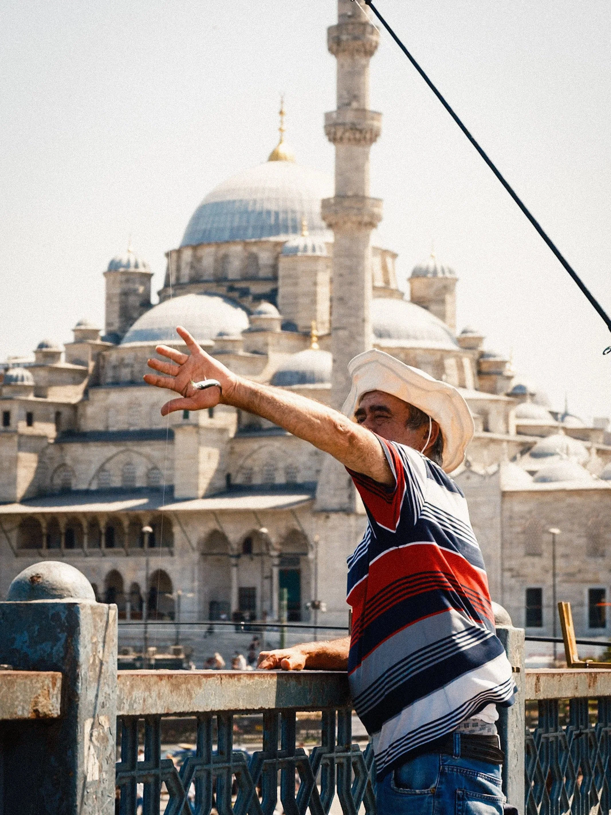 Man standing on the Galata Bridge and casting a fishing line with the domes and minarets of the New Mosque (Yeni Camii) rising behind him — classic Istanbul street photography capturing local fishing culture and iconic Ottoman architecture.