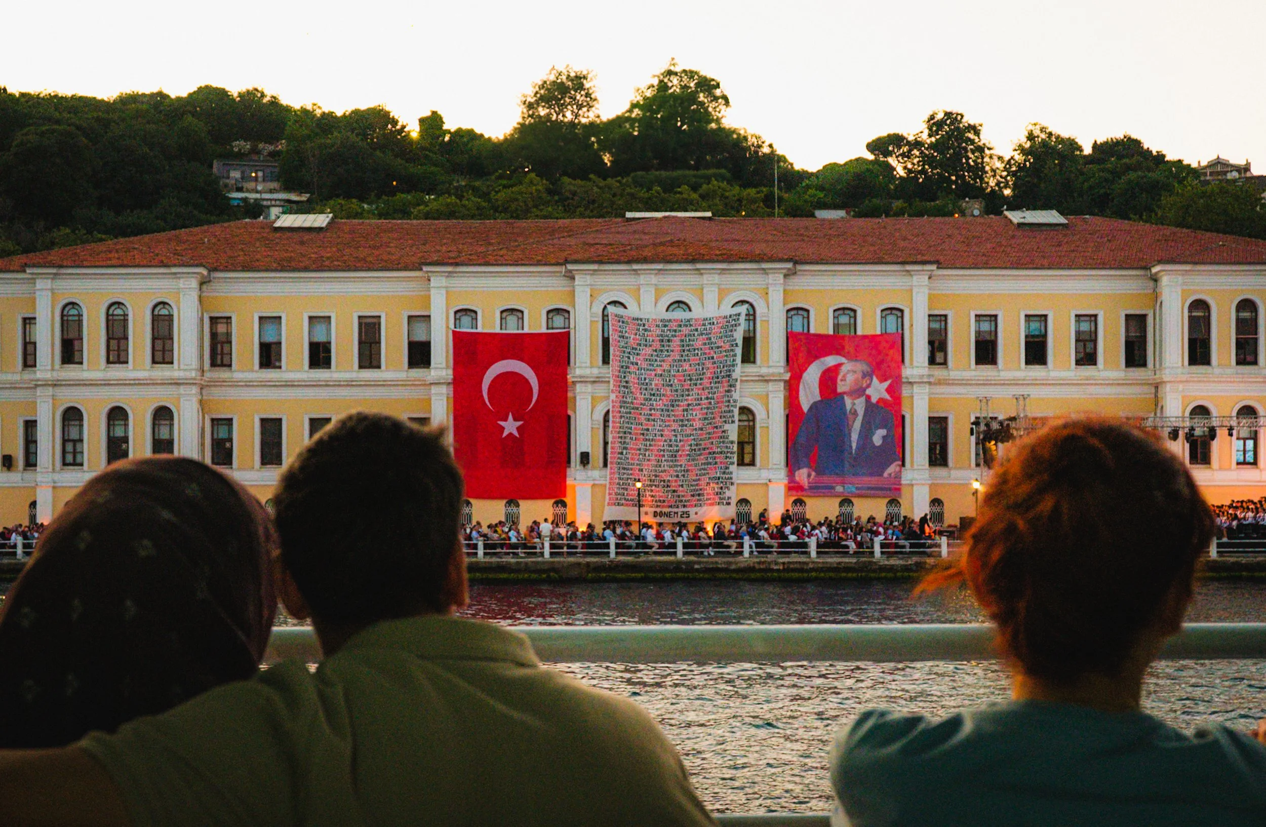 People sitting by the Bosphorus waterfront and watching a large Turkish flag and banners displayed on the façade of a historic building in Istanbul during sunset — atmospheric Istanbul street photography highlighting national celebrations and city sc