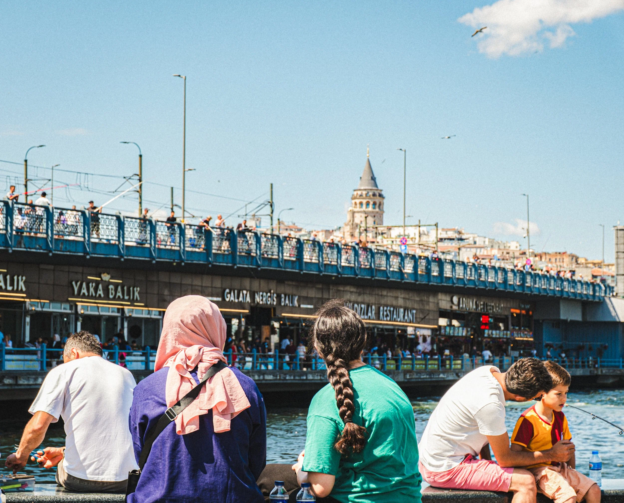 People sitting by the waterfront and looking toward the Galata Bridge and the Galata Tower rising over the Karaköy skyline in Istanbul — classic Istanbul street photography with a focus on everyday moments and iconic landmarks.