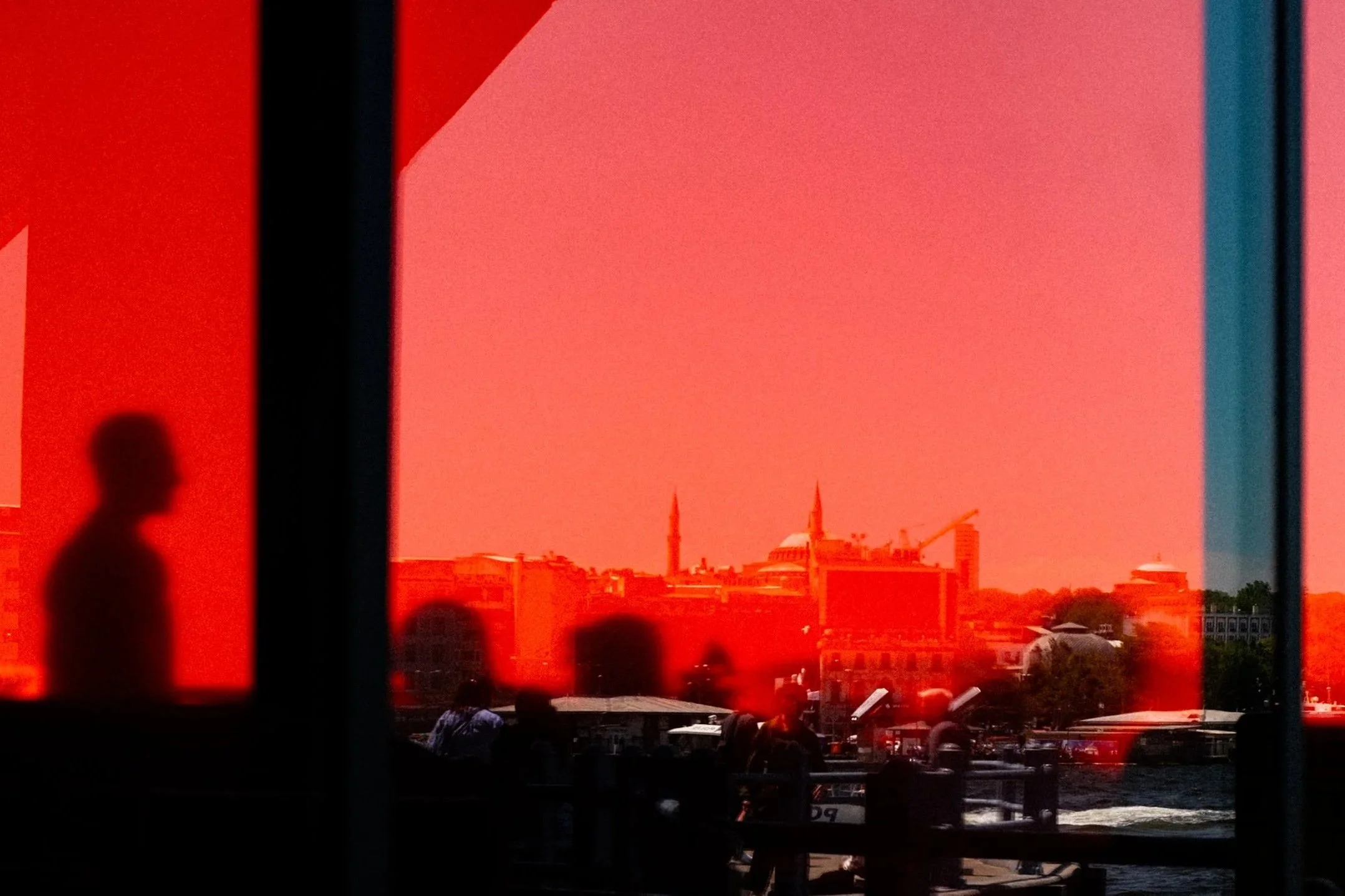 An abstract Istanbul street photography scene showing a silhouette reflected on bright red-tinted glass overlooking the Golden Horn. Through the colored surface, the city skyline, mosques, and waterfront activity appear distorted yet recognizable, bl