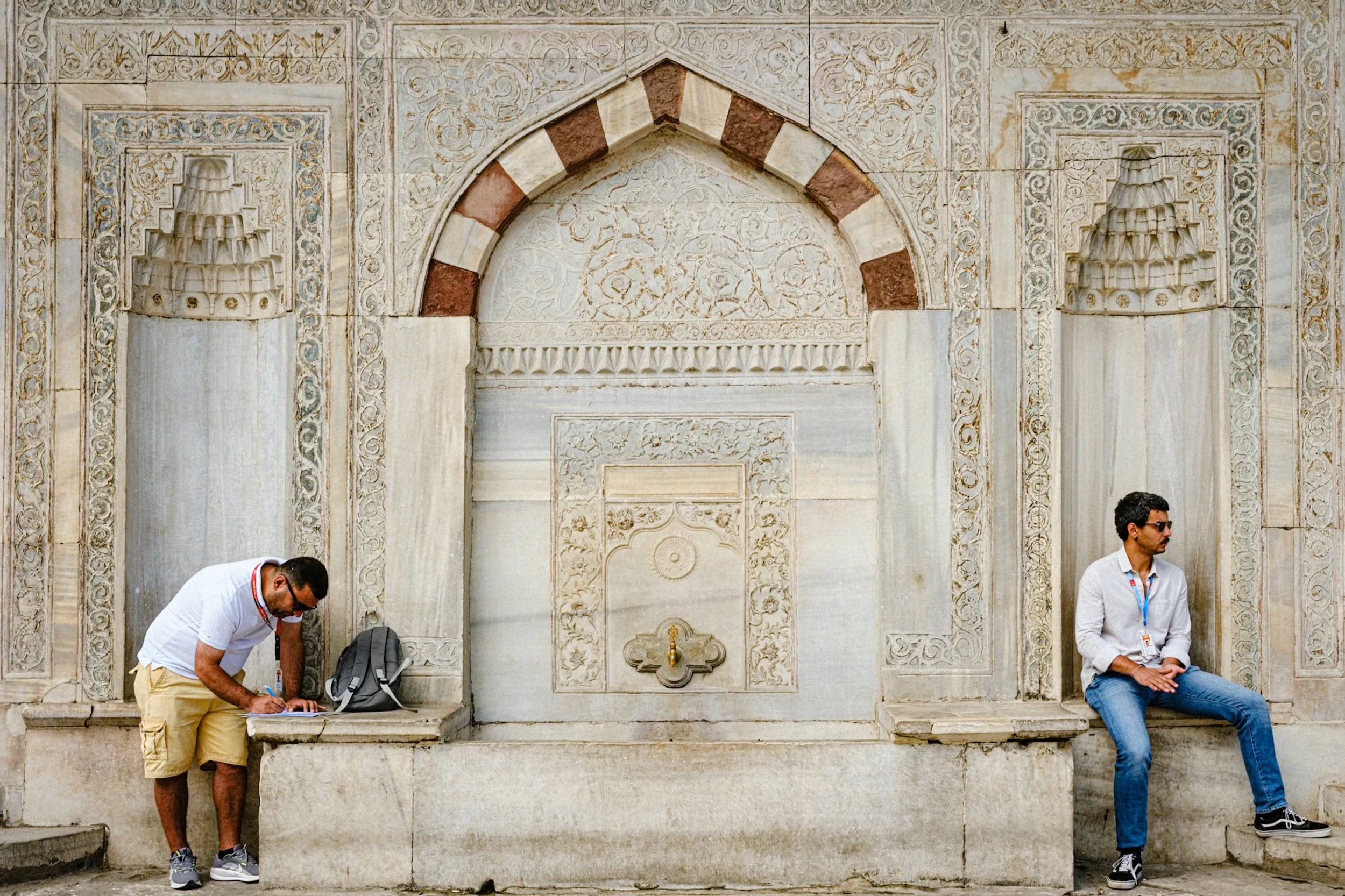Two men sitting near an ornate Ottoman-era stone fountain decorated with intricate carvings and arches in Istanbul — detailed Istanbul street photography highlighting historical architecture and quiet everyday scenes.