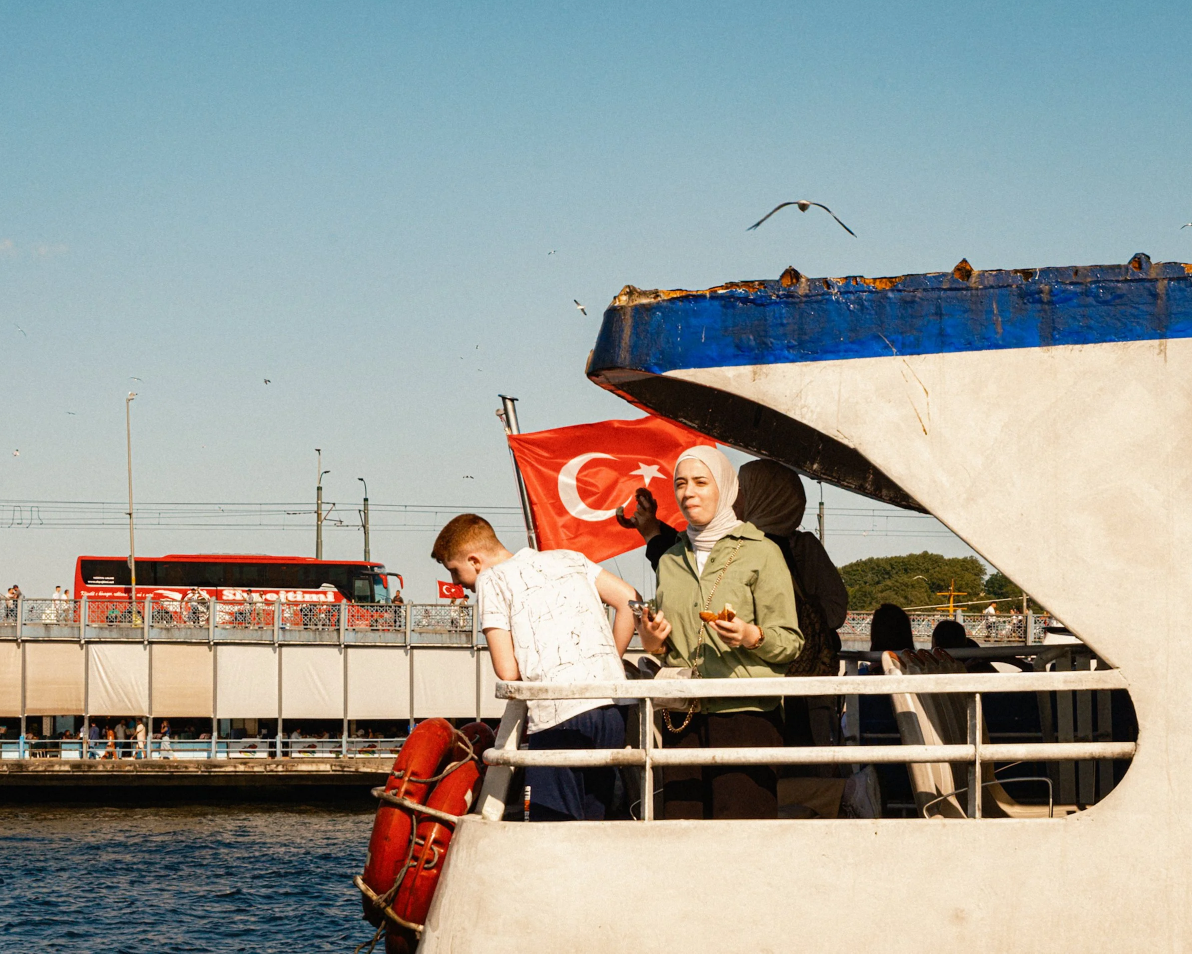 Passengers standing on a ferry deck with the Turkish flag waving behind them as the boat passes the Galata Bridge in Istanbul — dynamic Istanbul street photography capturing local transportation and daily scenes.