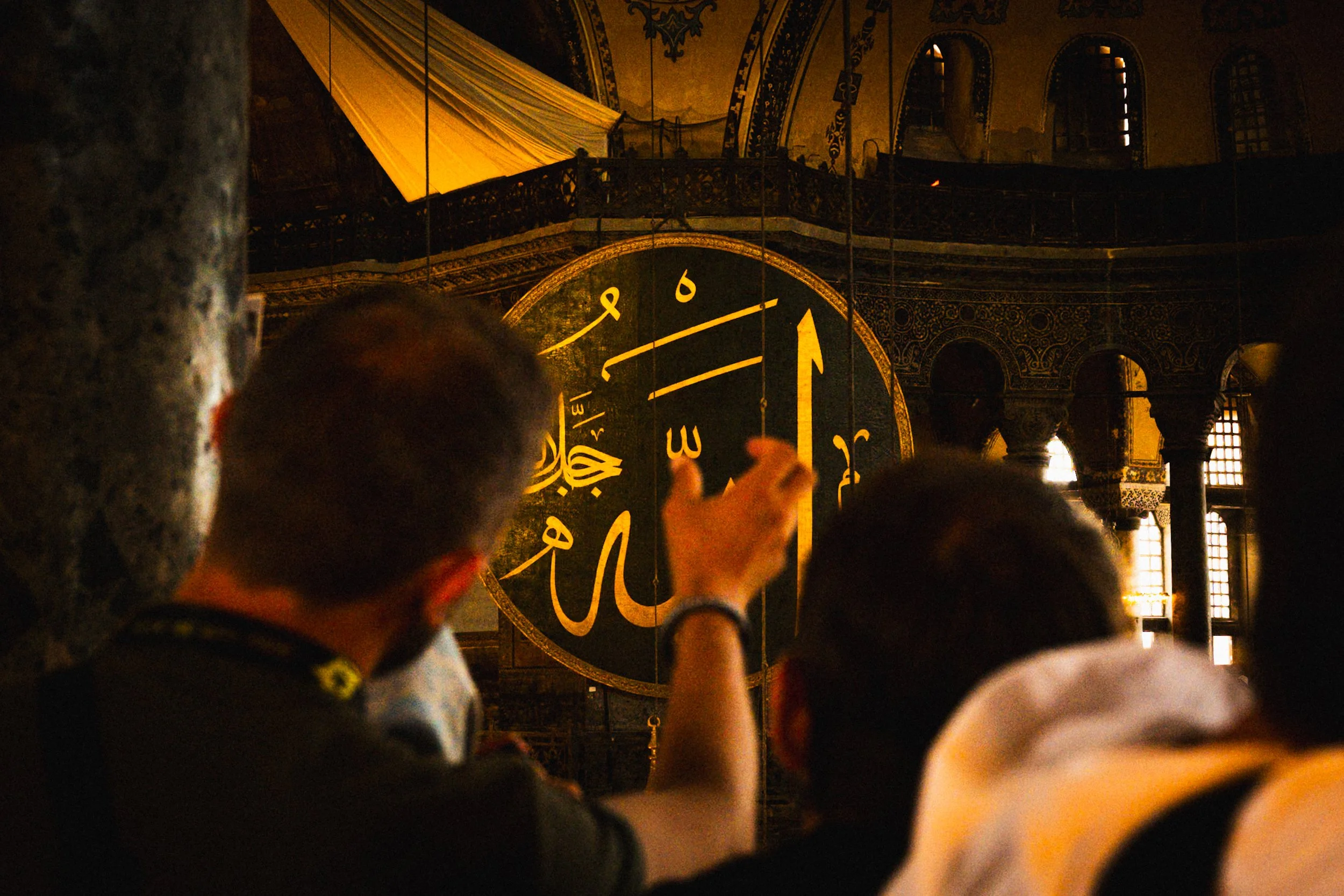 Visitors inside Hagia Sophia in Istanbul looking toward a large illuminated Arabic calligraphy medallion hanging beneath the dome — atmospheric Istanbul street and travel photography capturing historic Islamic–Byzantine architecture.