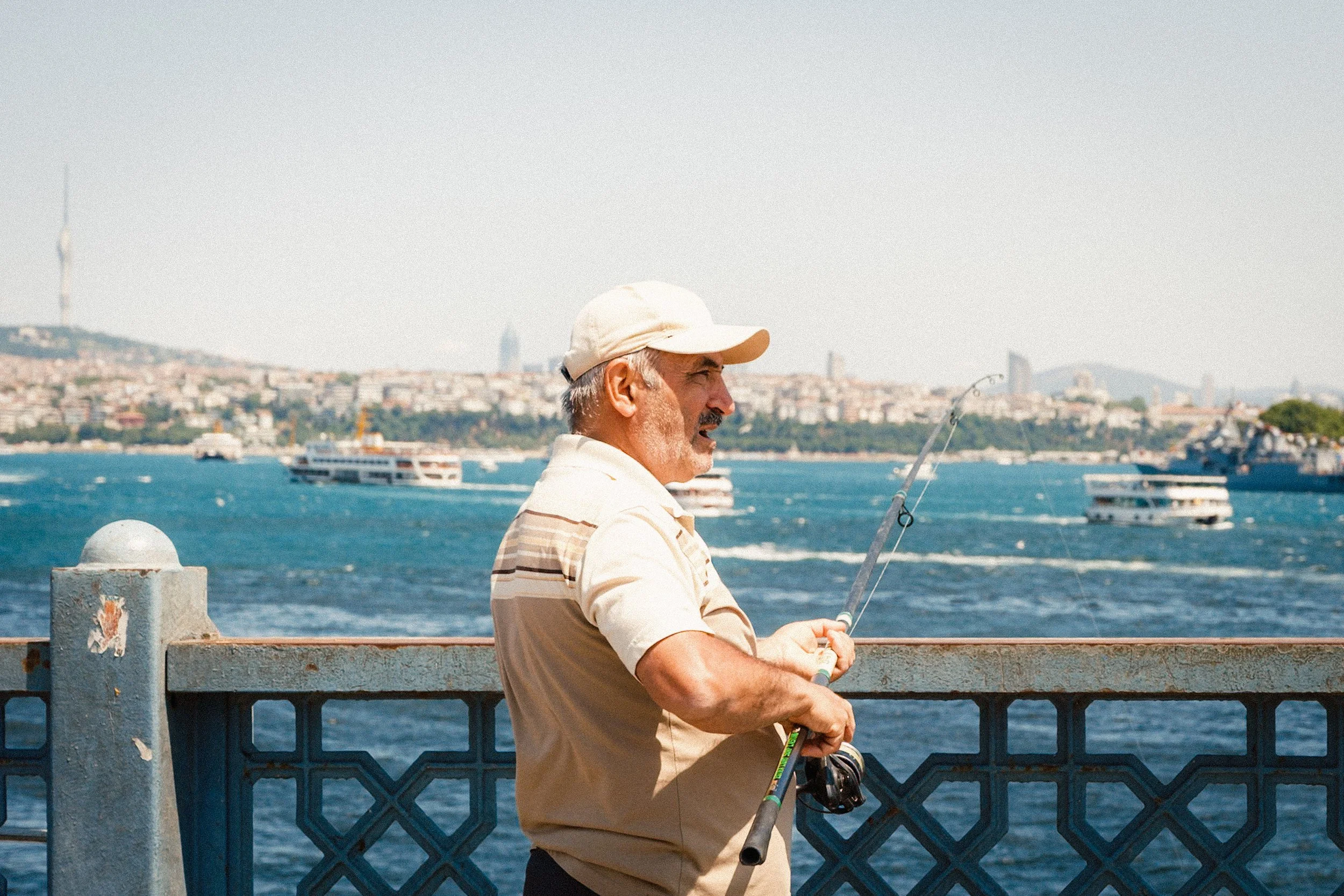 Man fishing from the Galata Bridge overlooking the Bosphorus, with ferries, hills, and the Istanbul skyline stretching across the horizon — classic Istanbul street photography focusing on peaceful daily routines.