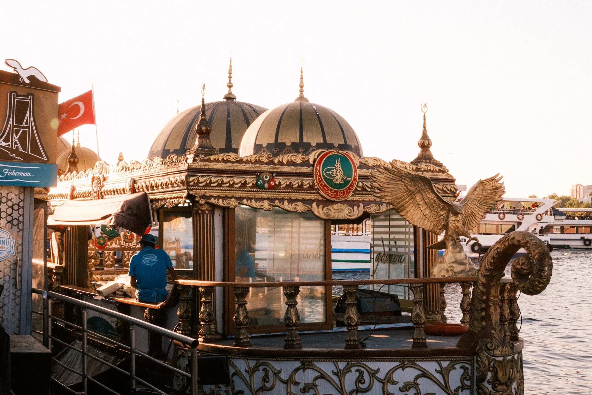 Traditional Ottoman-style fishing boat decorated with gold carvings, domes, and a winged figure, docked near the Eminönü waterfront in Istanbul during golden hour — vibrant Istanbul street and travel photography capturing local food culture.