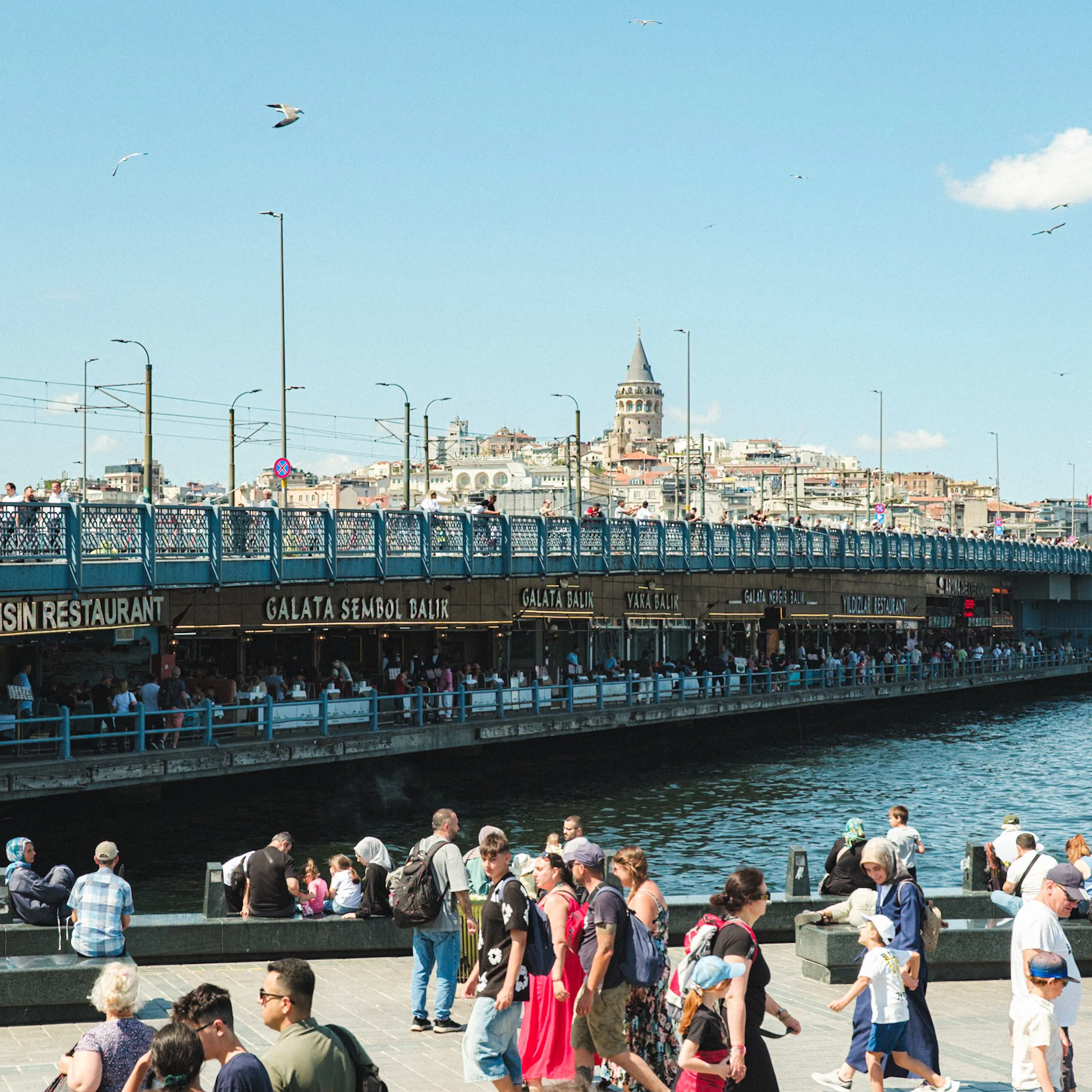 Crowds walking along the waterfront beneath the Galata Bridge in Istanbul, with restaurants lining the lower deck and the Galata Tower rising in the background — iconic Istanbul street photography scene.