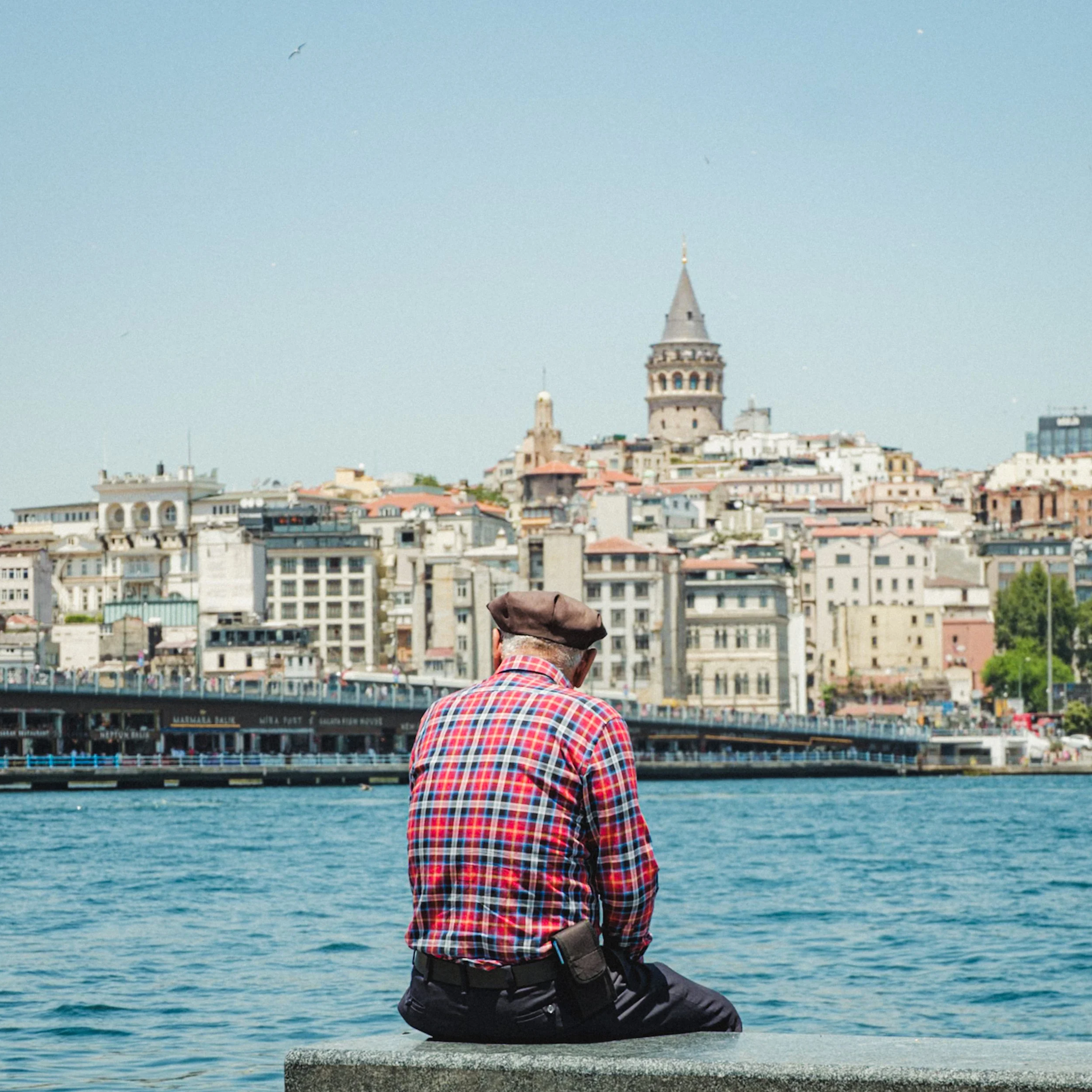 Elderly man in a plaid shirt sitting by the waterfront and looking toward the Galata Tower and the Beyoğlu skyline across the Golden Horn in Istanbul — atmospheric Istanbul street photography capturing quiet everyday moments.